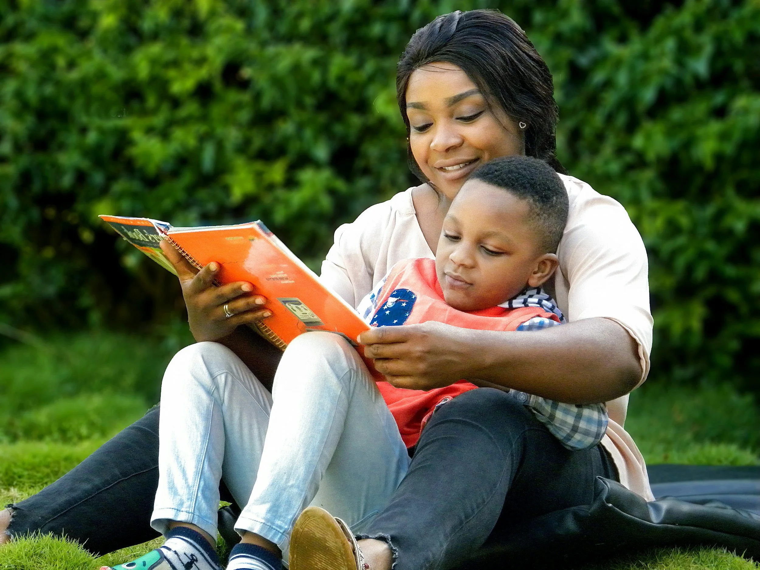 A woman with dark hair and earrings smiling while reading a book to a young boy outdoors on a grassy area, with green bushes in the background.
