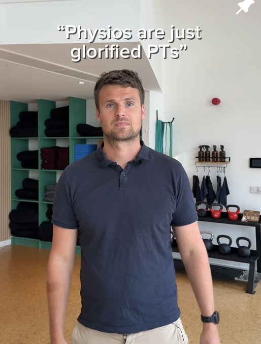 Man standing in a gym or fitness studio with workout equipment and shelves of towels in the background.