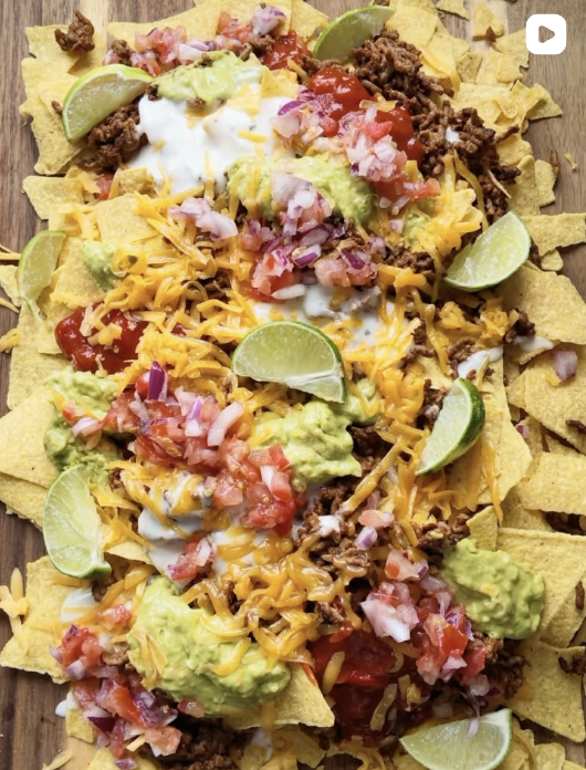 Loaded nachos with ground beef, shredded cheddar, diced tomatoes, guacamole, sour cream, chopped red onions, and lime wedges on tortilla chips.