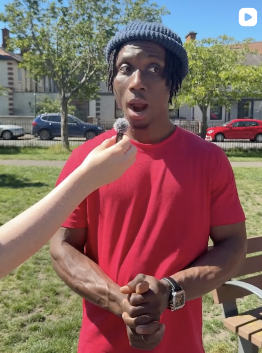 Young man in red t-shirt and beanie being interviewed outdoors on a sunny day, with trees, houses, and parked cars in the background.