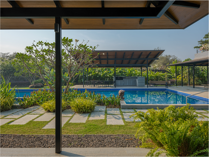 View of a modern backyard with a swimming pool, surrounded by lush greenery and patio furniture, seen from under a covered patio area. In Alibag.