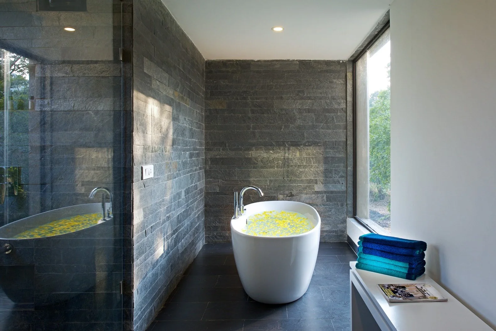 Modern bathroom with a stone wall, standalone bathtub filled with yellow flowers, glass shower enclosure, and folded blue towels on a counter.