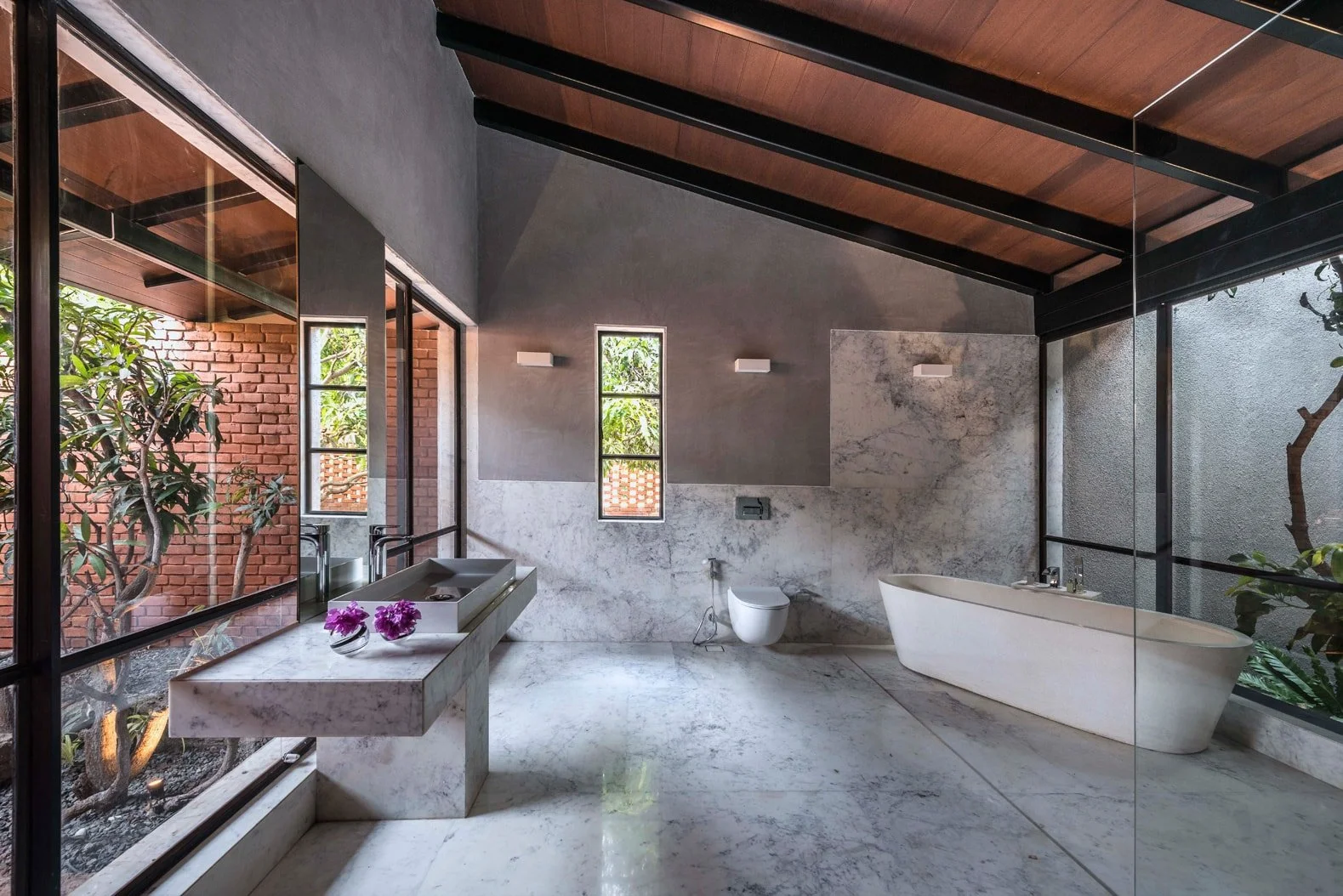 Modern bathroom with marble surfaces, freestanding bathtub, floating sink, and large windows showcasing outdoor greenery.