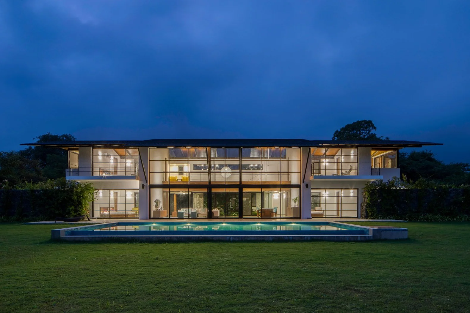 Modern two-story house with large windows and a swimming pool at night, surrounded by a lawn and trees.