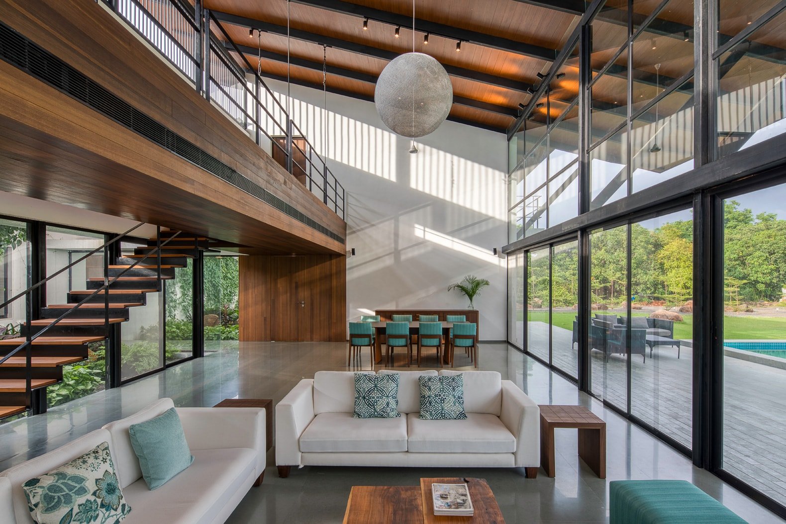 Modern living room with high ceilings, large glass windows, white sofas, wooden accents, floating staircase, dining table with turquoise chairs, and view of outdoor greenery.