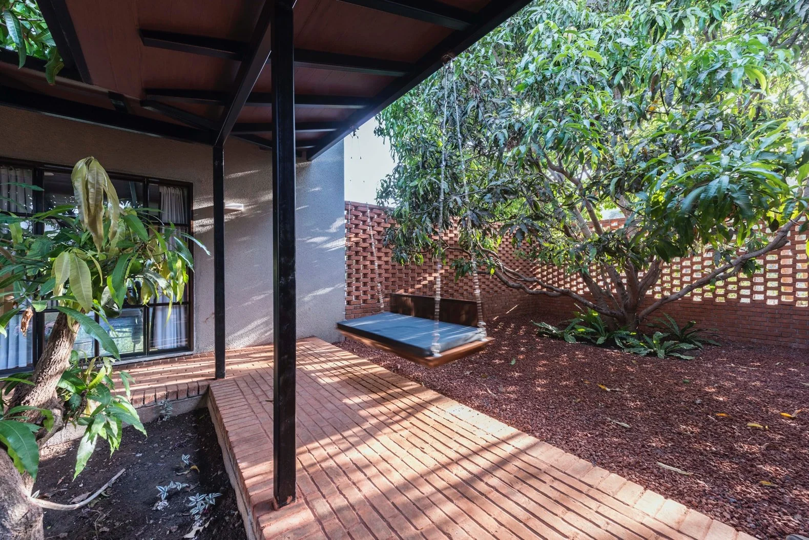 Outdoor patio with a wooden swing, brick flooring, metal canopy, and a large leafy tree in a garden setting.