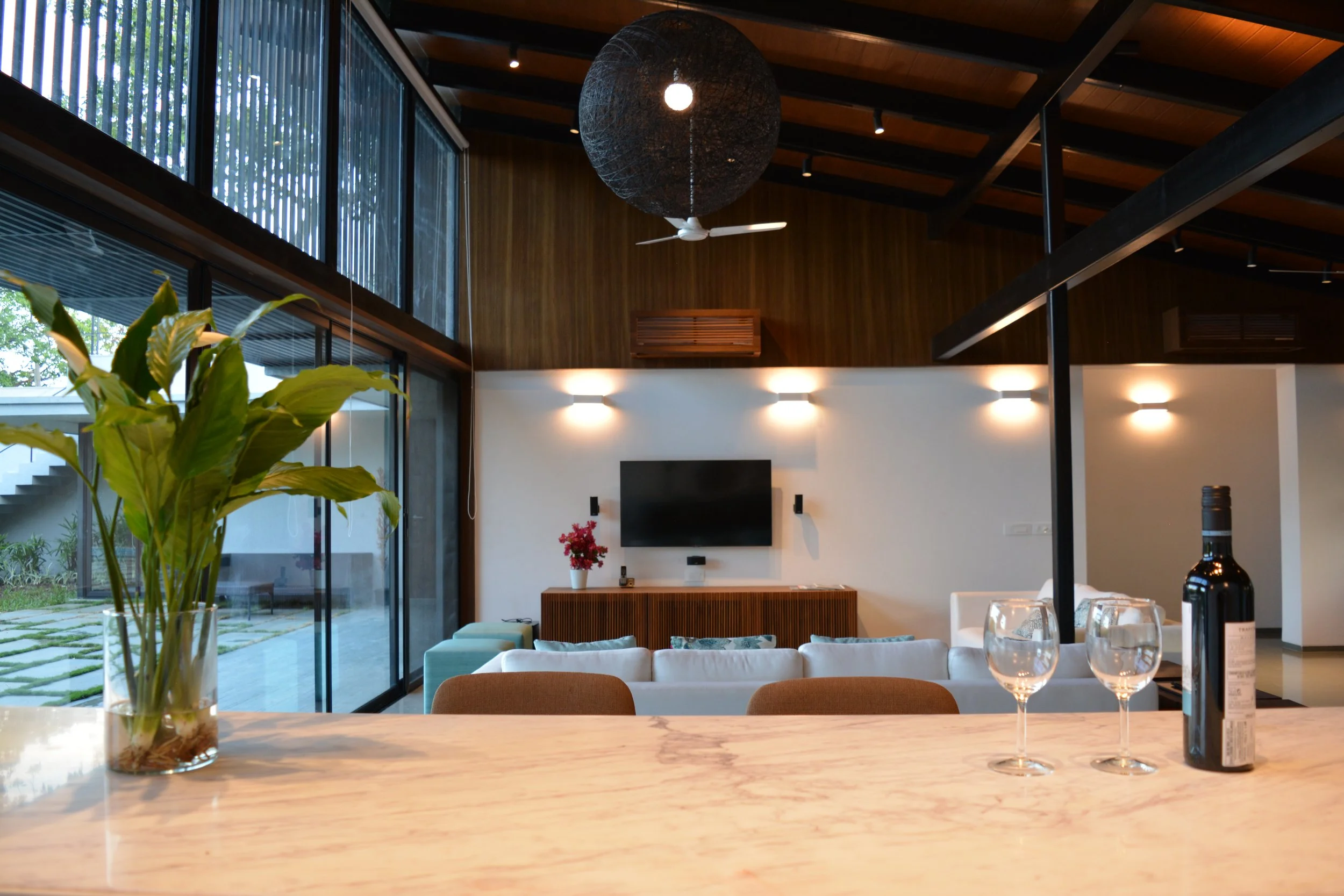 Modern living room with glass wall, ceiling fan, TV, sofa, and decorative lighting. Wine bottle and glasses on marble countertop, with a plant in the foreground.