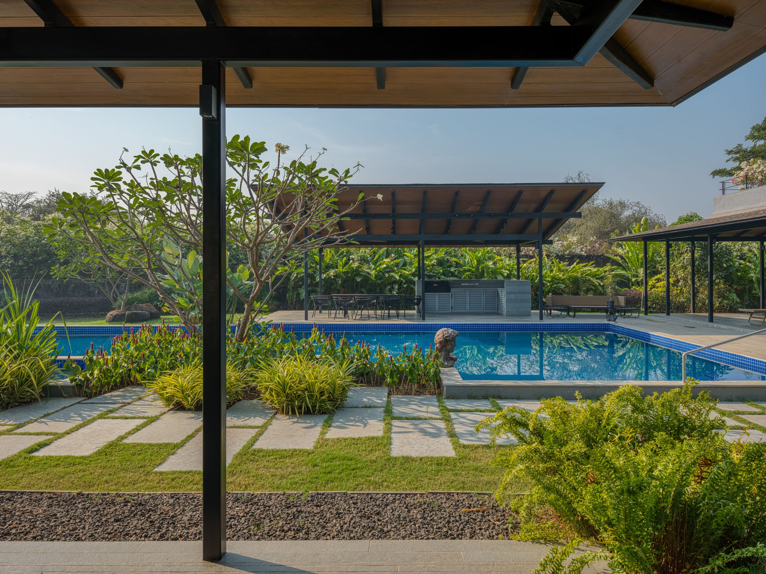 Outdoor swimming pool area with lush greenery, covered seating area, and lounge chairs, viewed from under a wooden roof.
