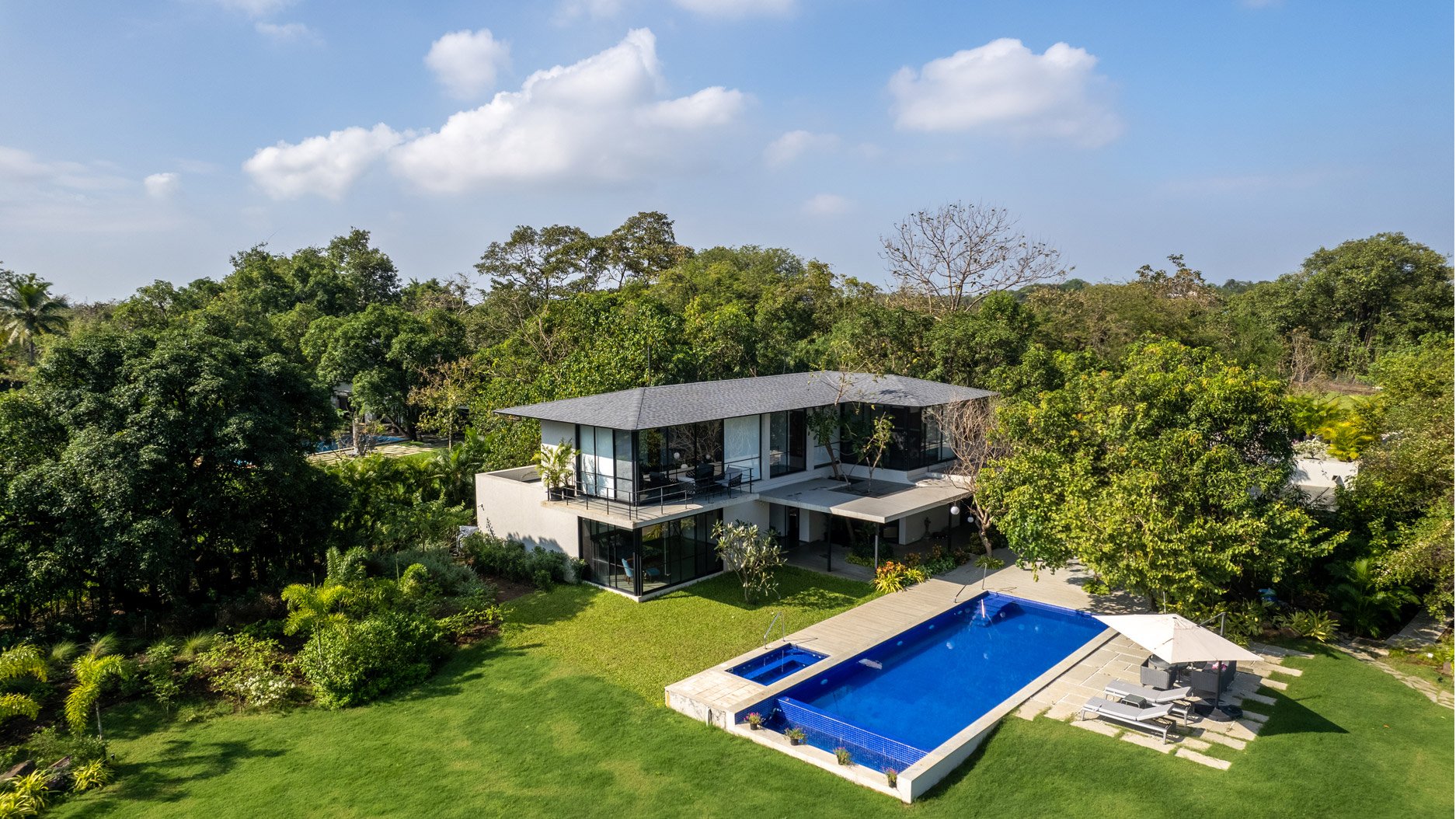 Modern house with large glass windows, surrounded by lush greenery and trees, featuring a swimming pool with lounge chairs and a shaded area in the backyard.
