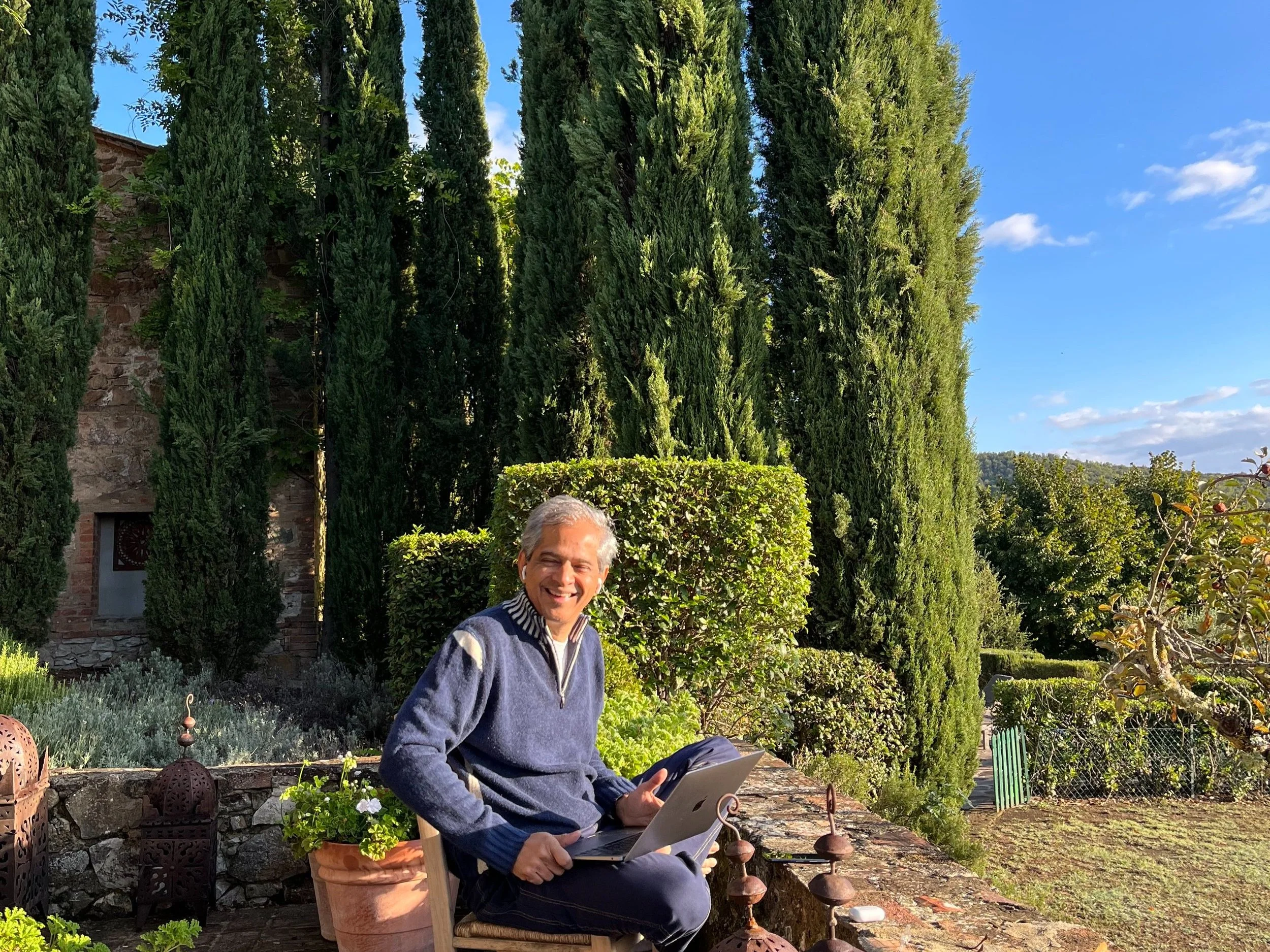 Person sitting outdoors with laptop, surrounded by tall trees and greenery, smiling on sunny day.