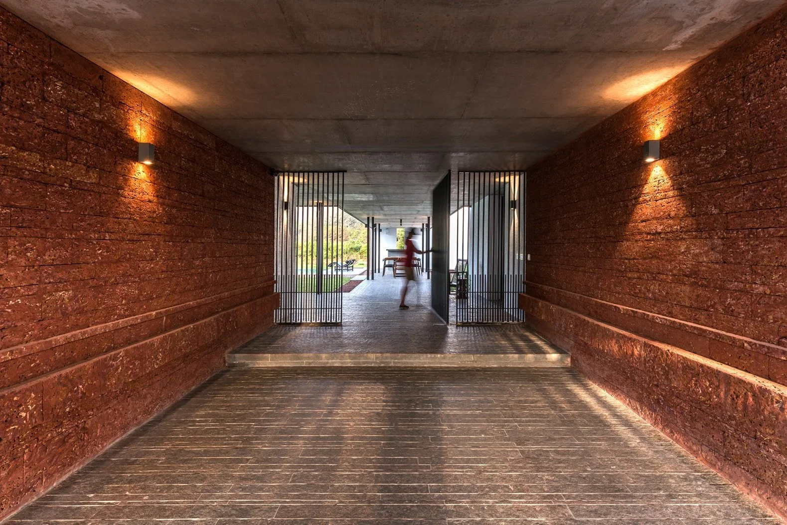 Long hallway with modern, textured red walls and ceiling lighting, leading to an open area with a blurred figure walking.