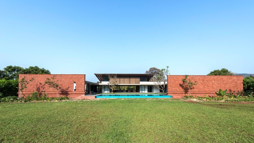 Modern house with red brick walls, large windows, and a swimming pool, surrounded by a green lawn and trees.