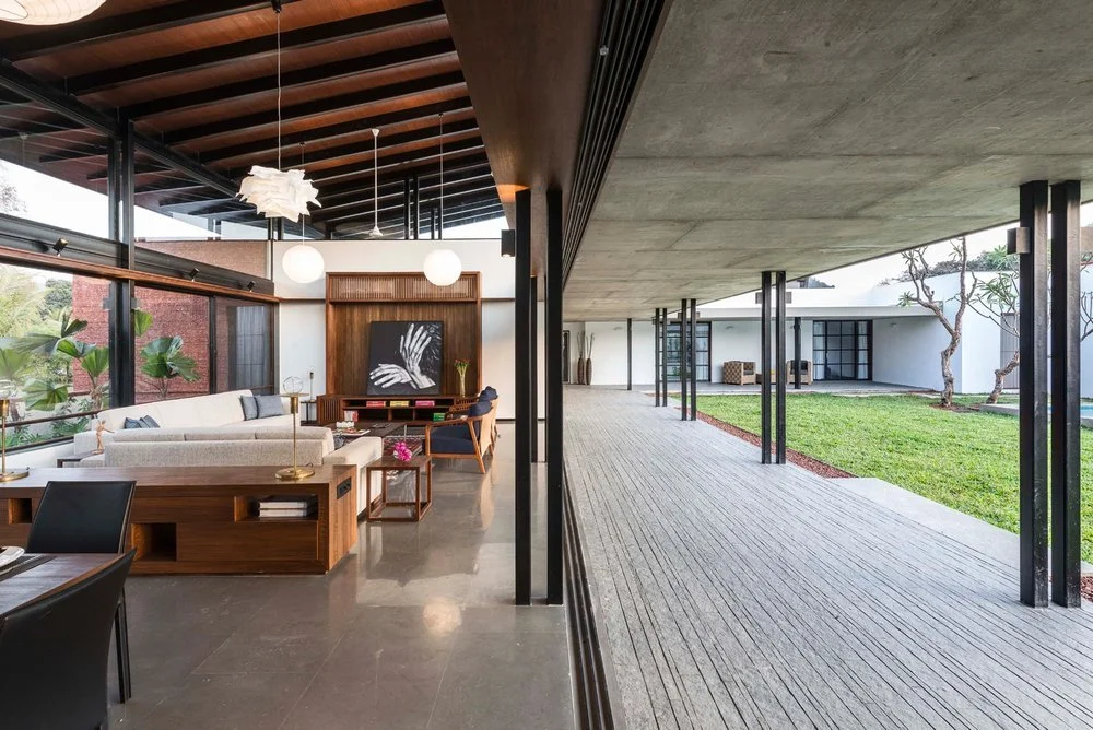 Modern living room with large windows, a sectional sofa, wooden furniture, and contemporary lighting. Adjacent outdoor walkway and green lawn visible.