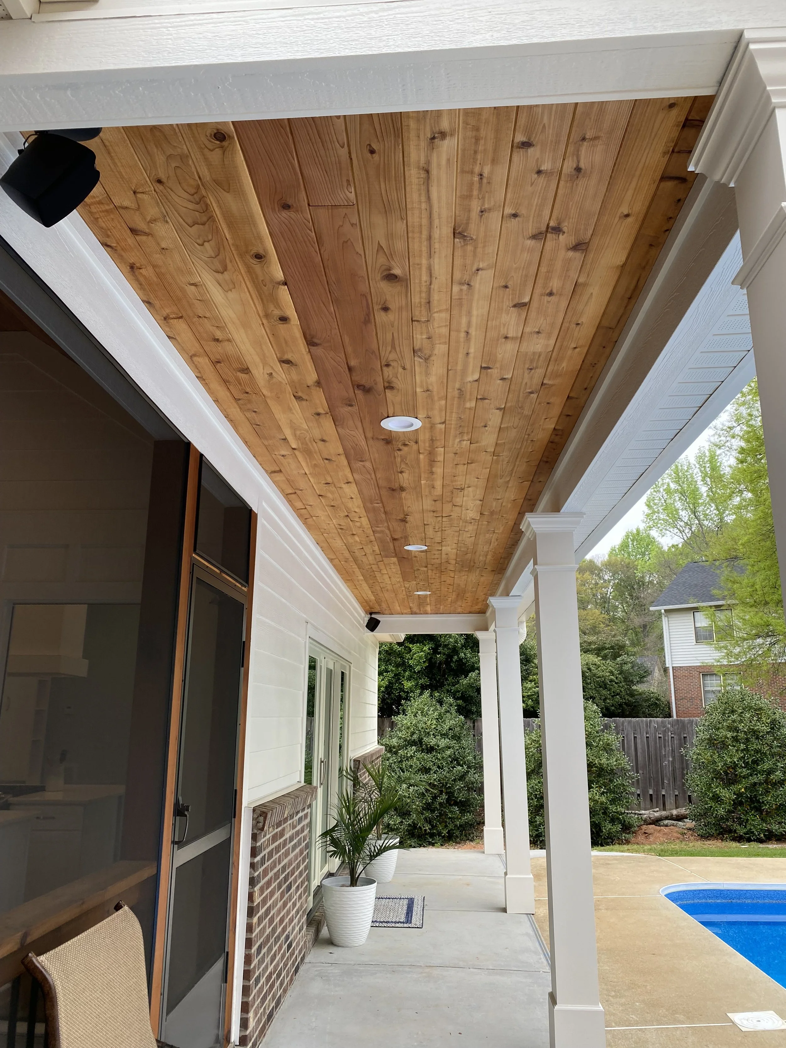Detached pool house front porch cedar tongue and groove ceiling with recessed can lights and outdoor speakers beside in ground pool.