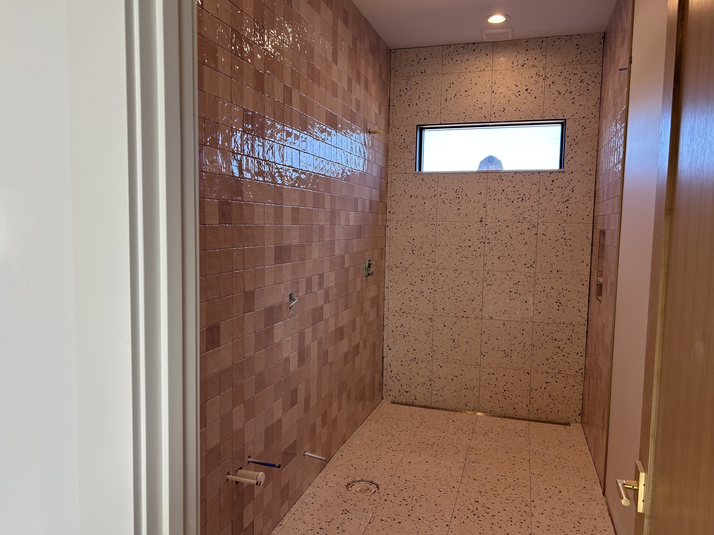 4x4 pink square tiles installed on bathroom wall to ceiling. 16"x16" real terrazzo tiles installed on floor and back wall of bathroom and shower. Curbless shower entry with gold trench drain against wall.