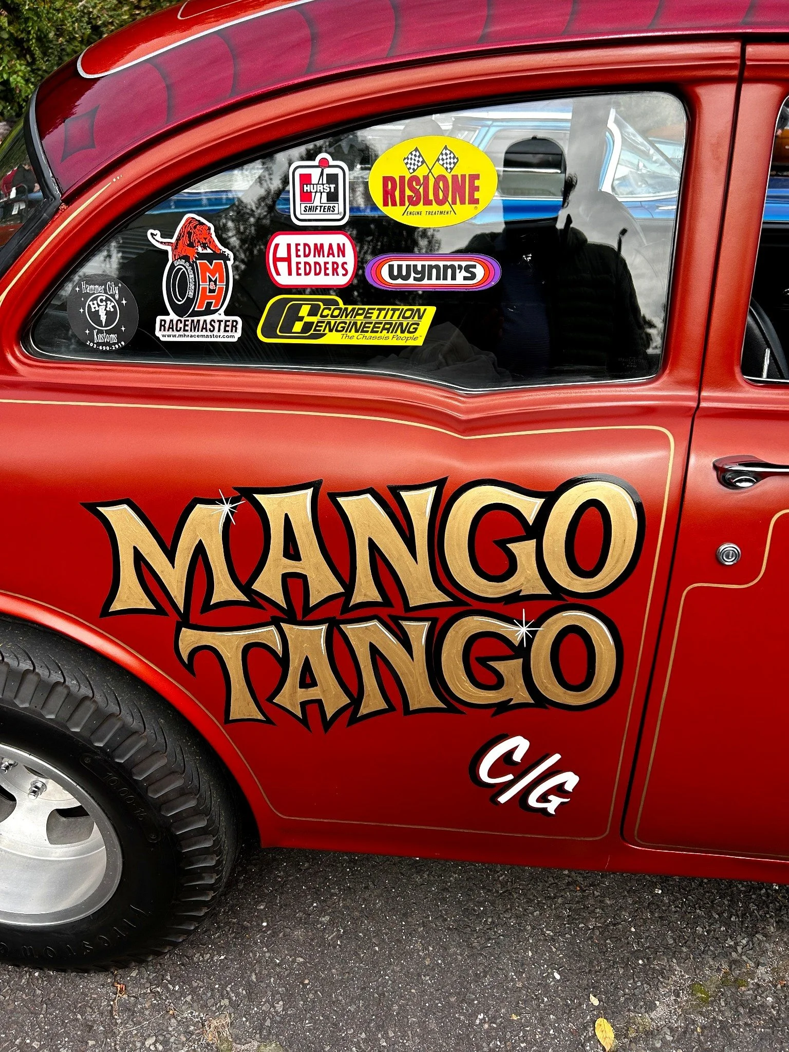 Red hot rod with gold hand-lettered "Mango Tango" on the door and a quarter window full of vintage racing decals.