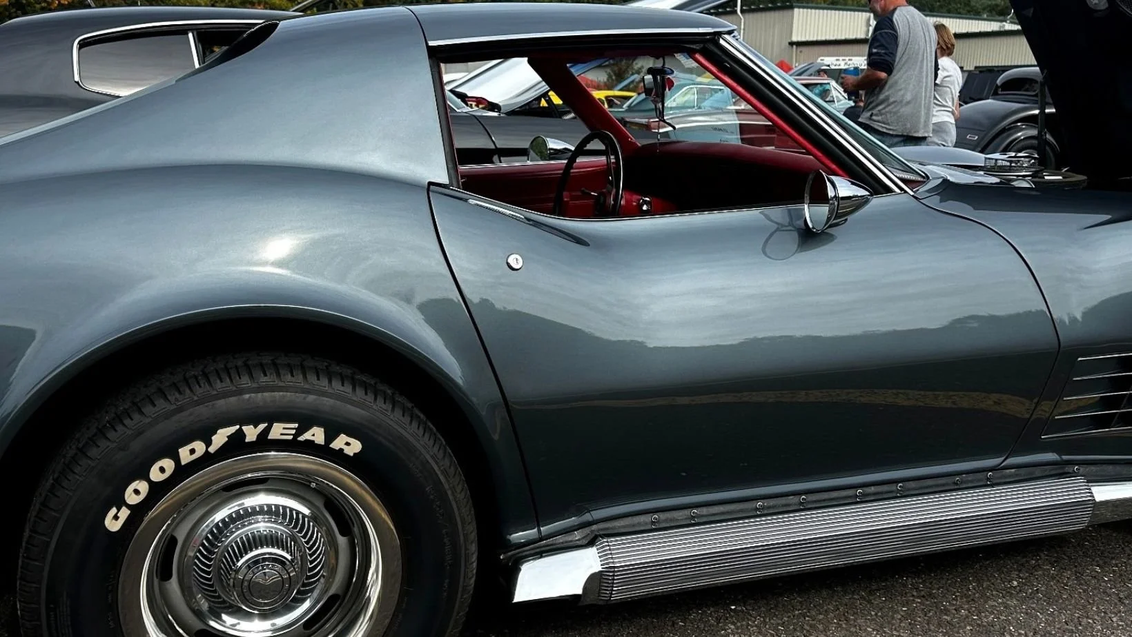 Side view of the gray C3 Corvette at the show, showing its red interior, chrome side mirror, side-exhaust trim along the rocker panel, and a Goodyear tire with raised white lettering.