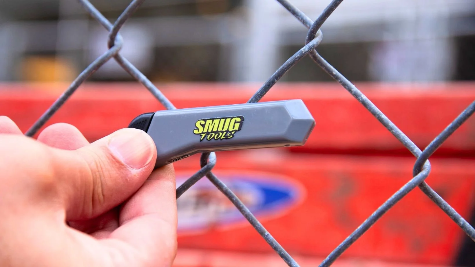 smug tools mini box cutter with blade retracted being held in a hand against a chain link fence