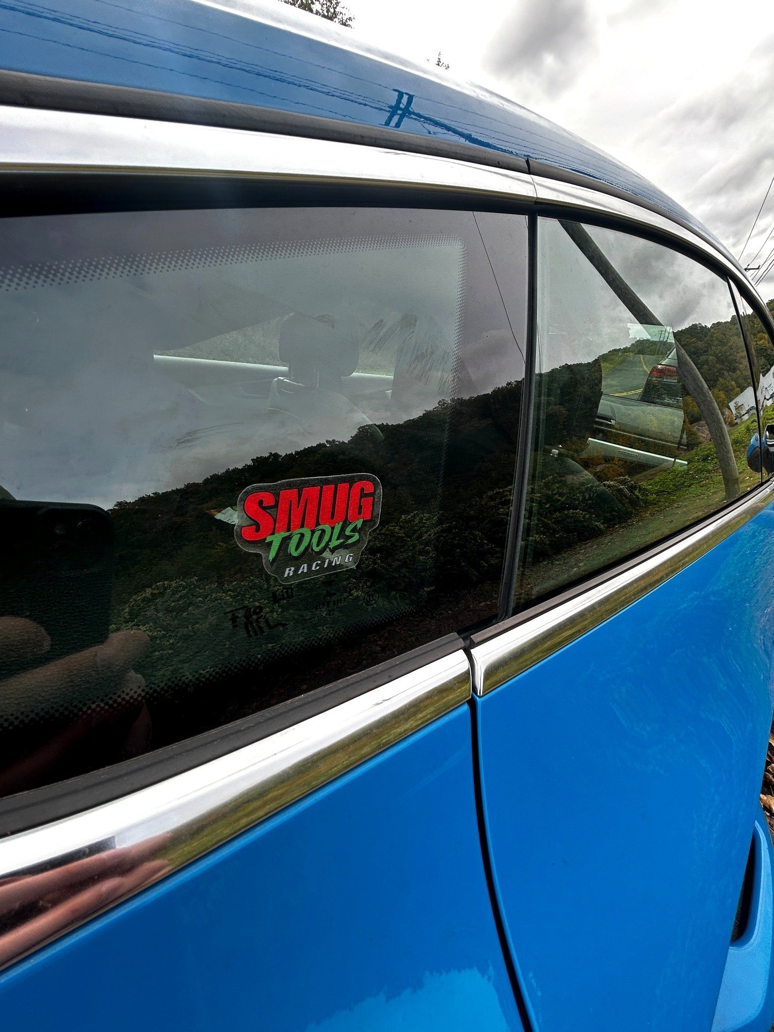Smug Tools Racing sticker on the rear quarter window of a bright blue car. The logo has "SMUG" in red, "TOOLS" in green, and "RACING" in white on a dark background. Trees and a hillside road are reflected in the glass.