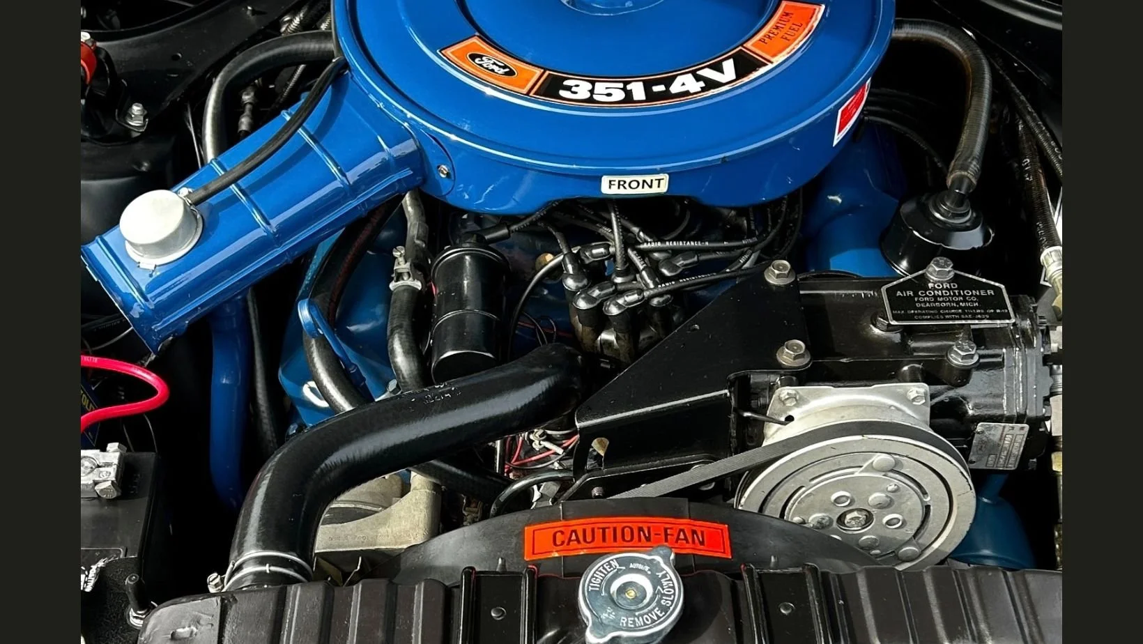 Close-up of a restored Ford 351-4V engine bay with a blue air cleaner bearing an orange "351-4V" decal, a black air conditioning compressor, and a red "Caution-Fan" warning sticker above the radiator cap.