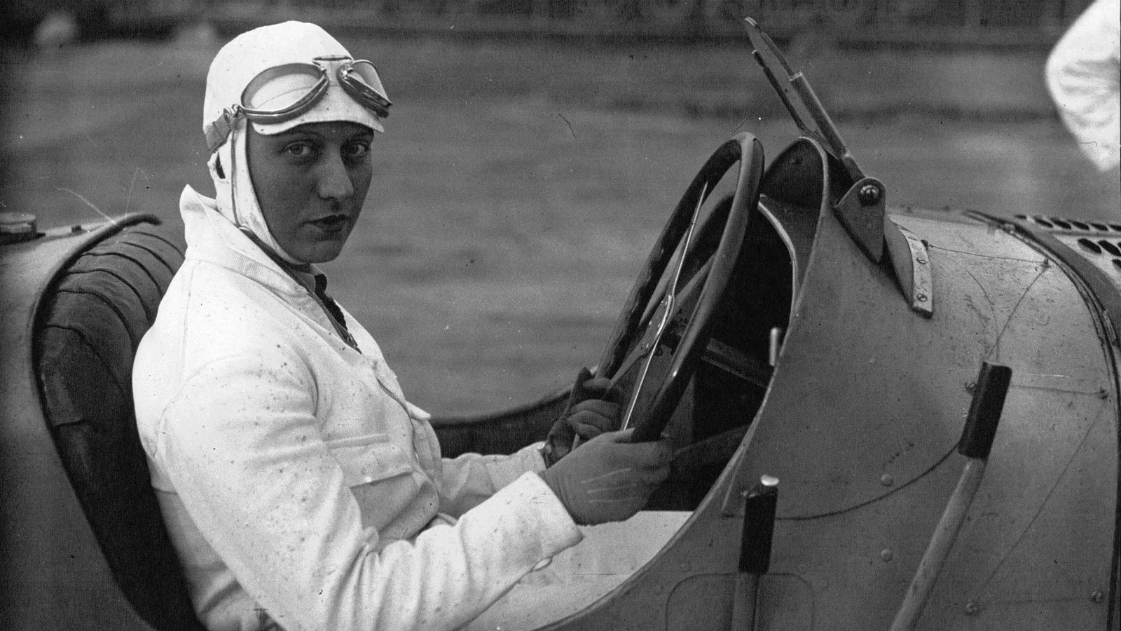 Hellé Nice wearing goggles and a racing suit while seated in a racing car at Montlhéry in 1929.