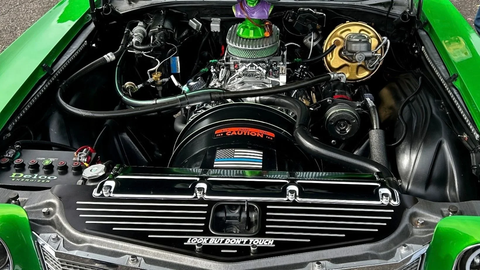 Detailed engine bay of a green classic car with a black finned radiator cover reading "Look But Don't Touch" and a small character bust on the air cleaner.
