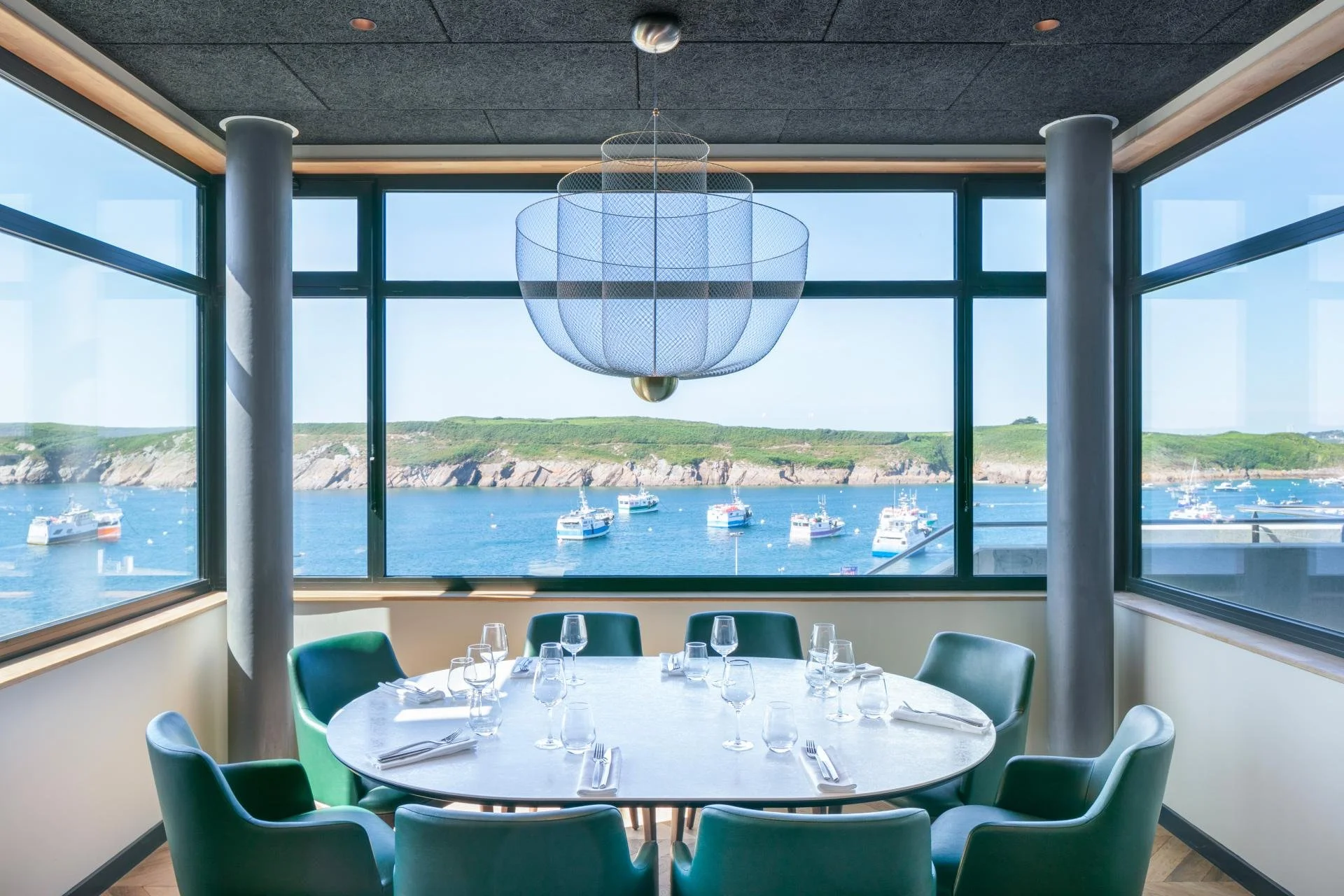 Salle à manger avec vue sur la mer et des bateaux, décoration moderne, grande fenêtre panoramique, chandelier design suspendu.