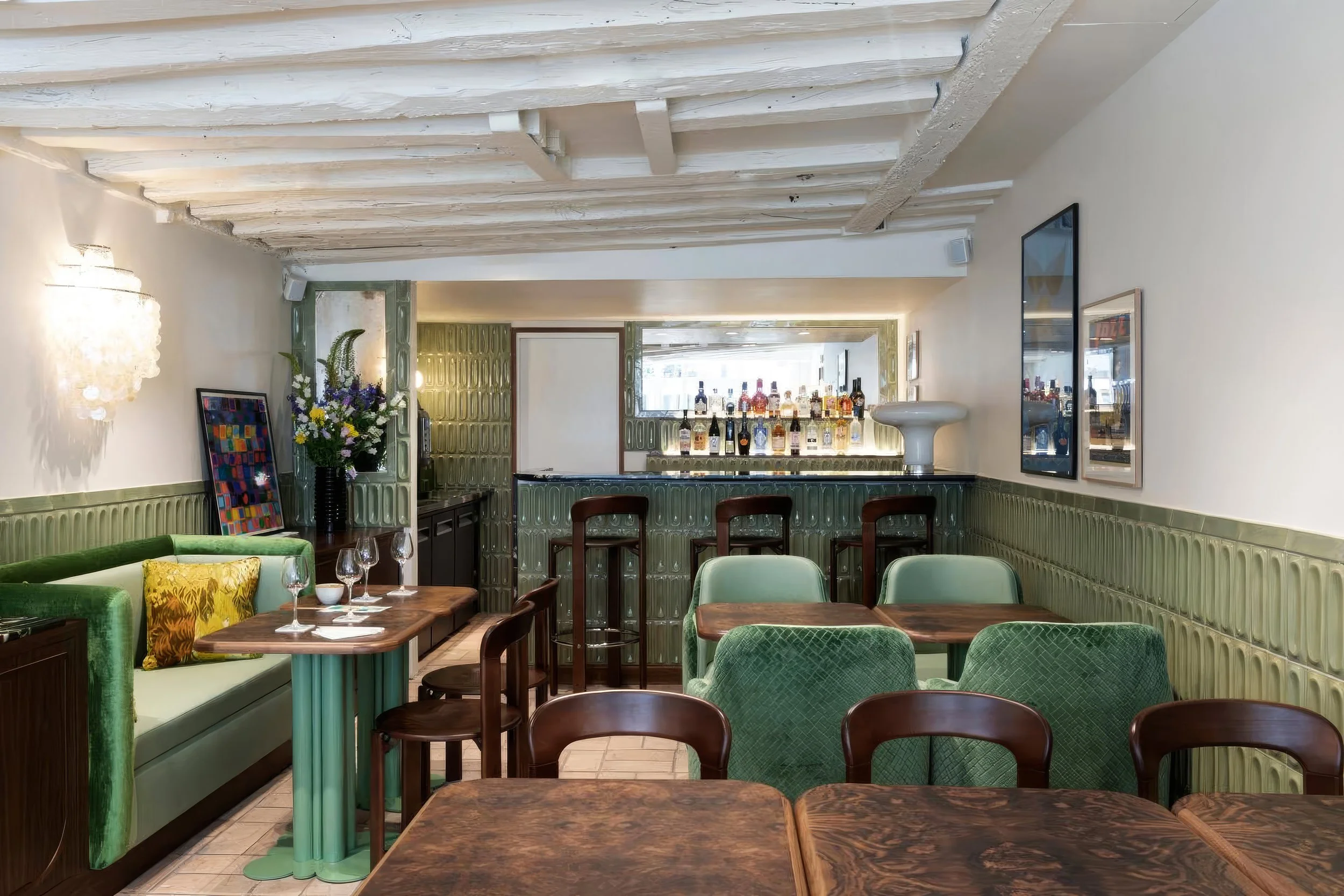 Intérieur d'un restaurant avec tables en bois, chaises en vert, décoration florale et exposition de bouteilles derrière un bar.