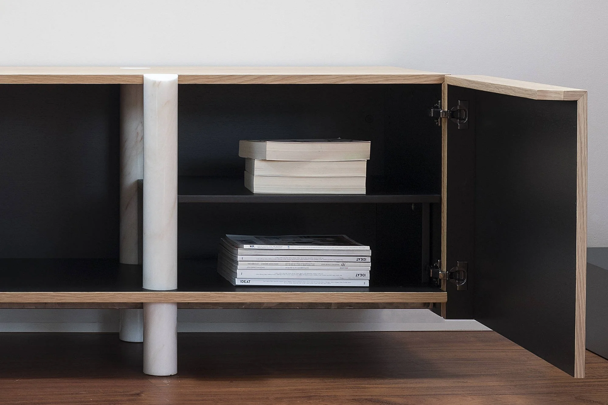 Une bibliothèque moderne en bois clair avec une porte ouverte, contenant des livres et des magazines, sur un sol en bois foncé.