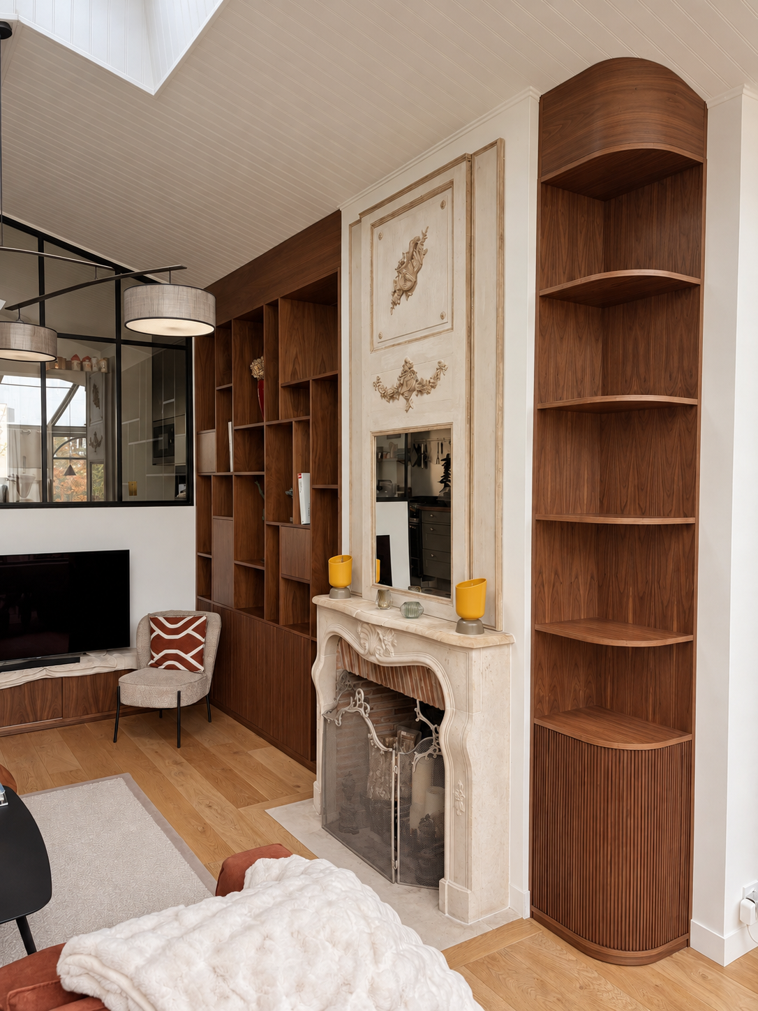 Intérieur d'un salon avec cheminée en marbre blanc, étagères en bois, une télévision, une chaise avec coussin kaki, et un plafond en bois peint en blanc.