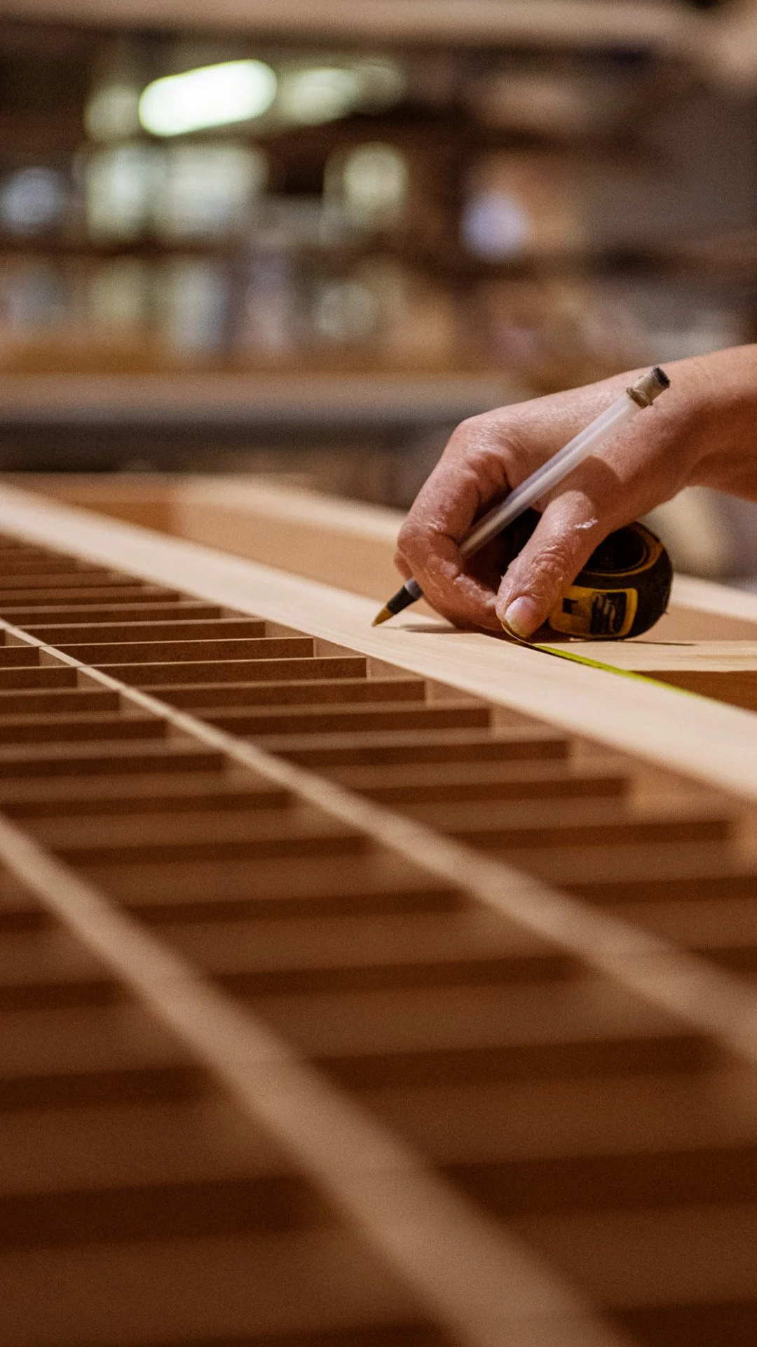 Une main mesure la longueur d'une planche en utilisant un ruban à mesurer dans un atelier de fabrication de meubles ou de guitare, avec une grille en bois en dessous.