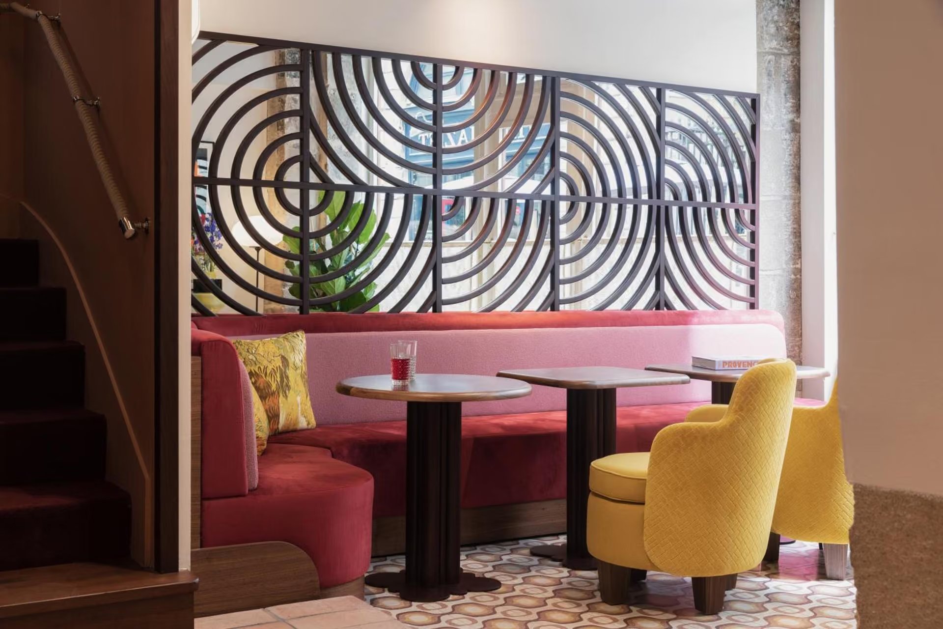 Coin salon avec banquette rose, fauteuil jaune, tables en bois, séparation en métal avec motifs en arc, plantes vertes, vin dans un verre, livres sur une table.