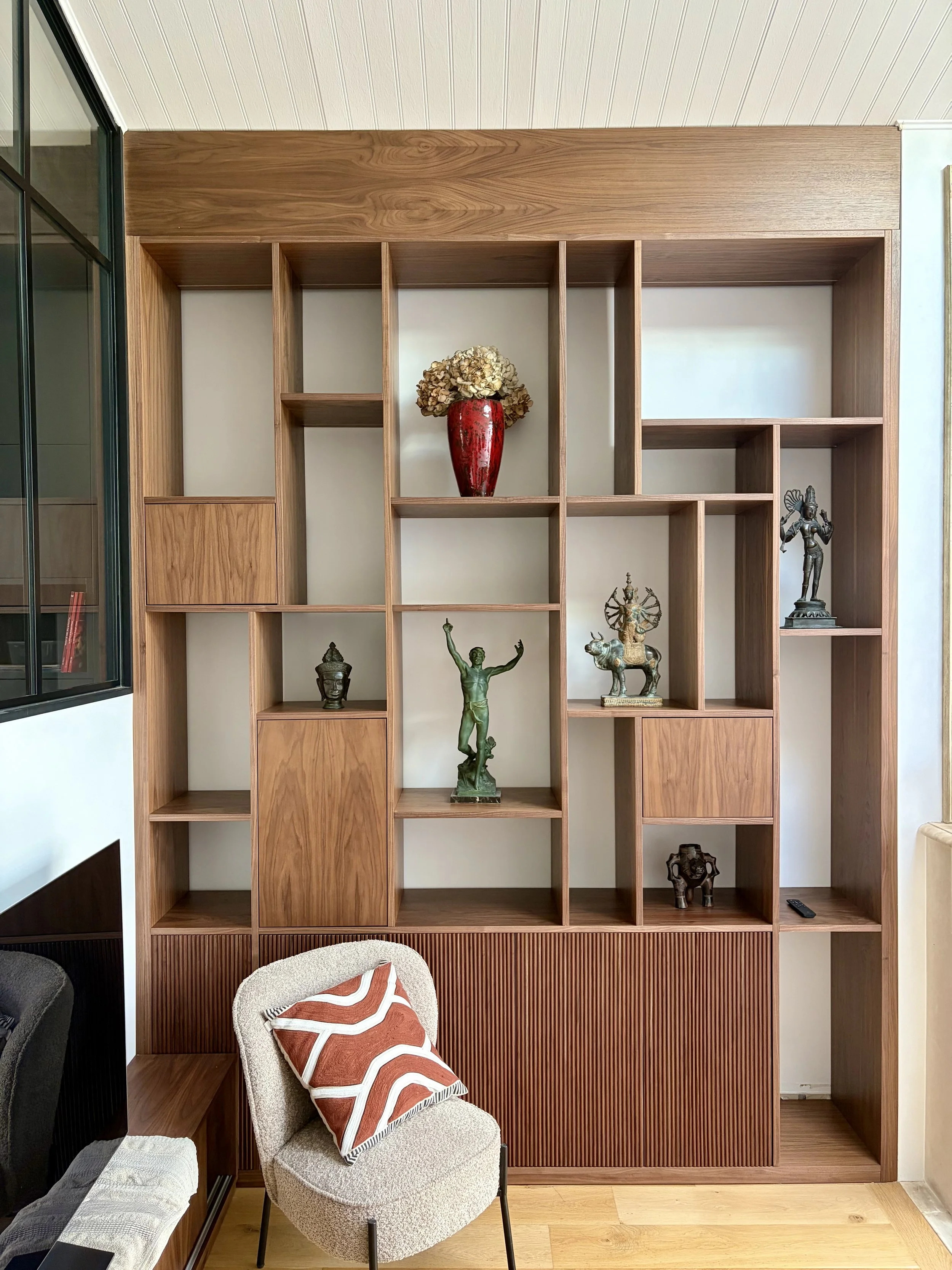 Une étagère en bois avec plusieurs compartiments contenant des statues indiennes, dont une grande en son centre, et des objets décoratifs. En bas, un fauteuil avec un coussin à motif géométrique rouge et blanc.
