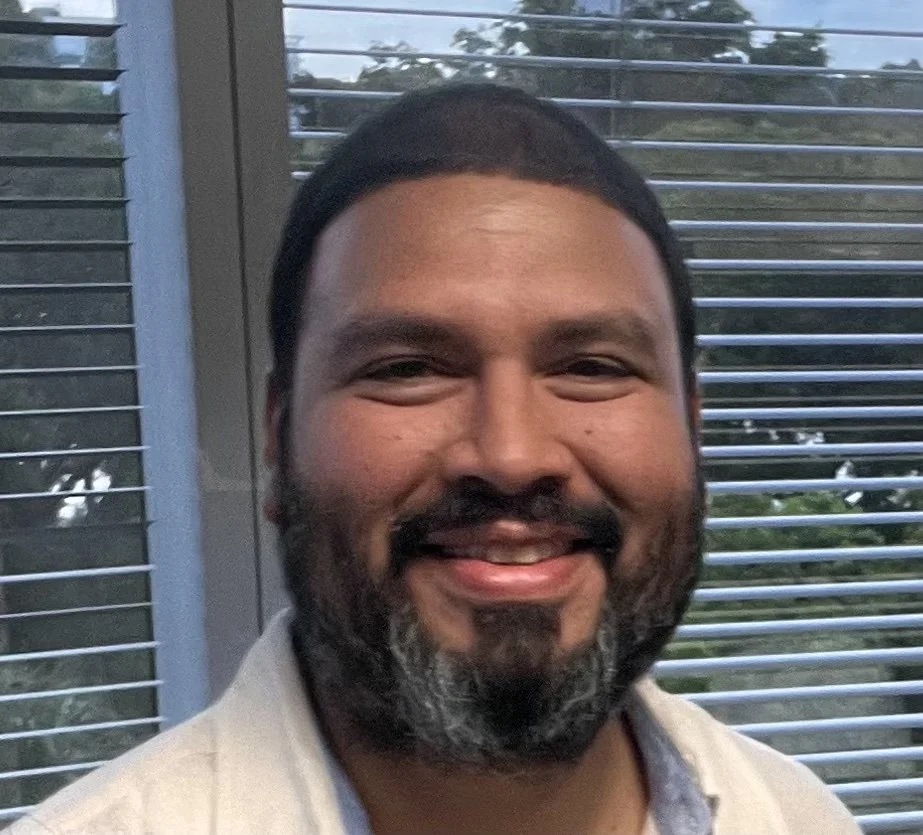A man with a beard and mustache smiling, standing in front of a window with blinds, outdoors trees visible outside.