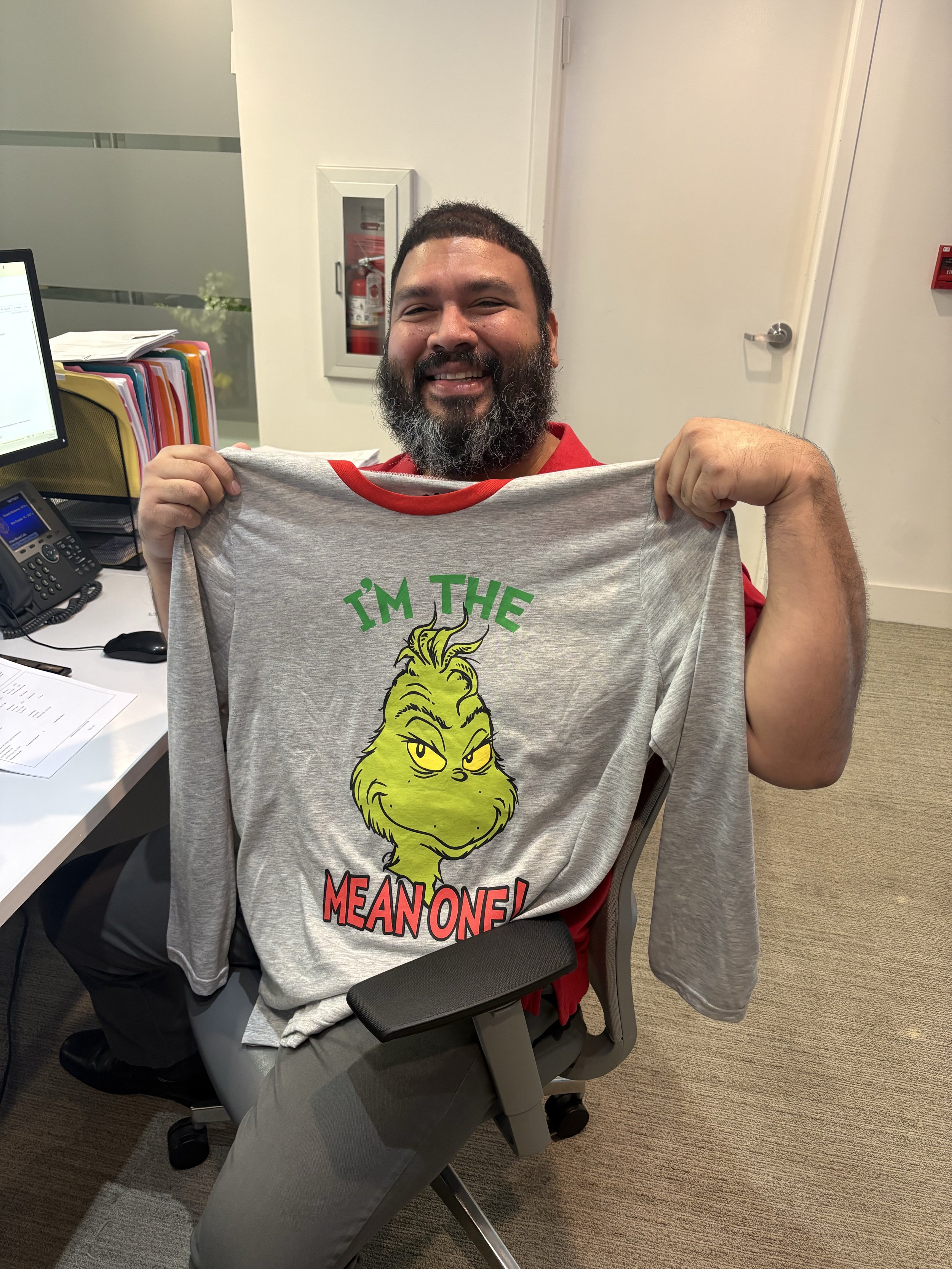 A man with a beard smiling and holding a Grinch-themed T-shirt. The T-shirt features the Grinch's face and the words 'I'm the mean one!'