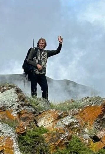 A man standing on a rocky mountain top, waving with his right hand, wearing a backpack, outdoor gear, and smiling.