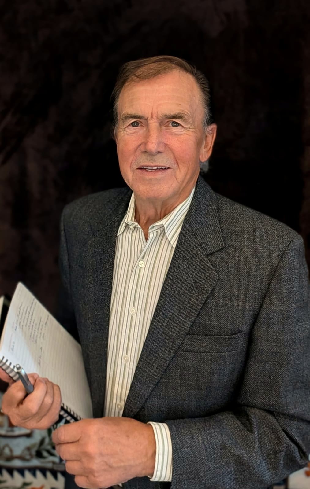 An elderly man holding a pen and notebook, wearing a gray blazer and a striped shirt, standing against a dark background.
