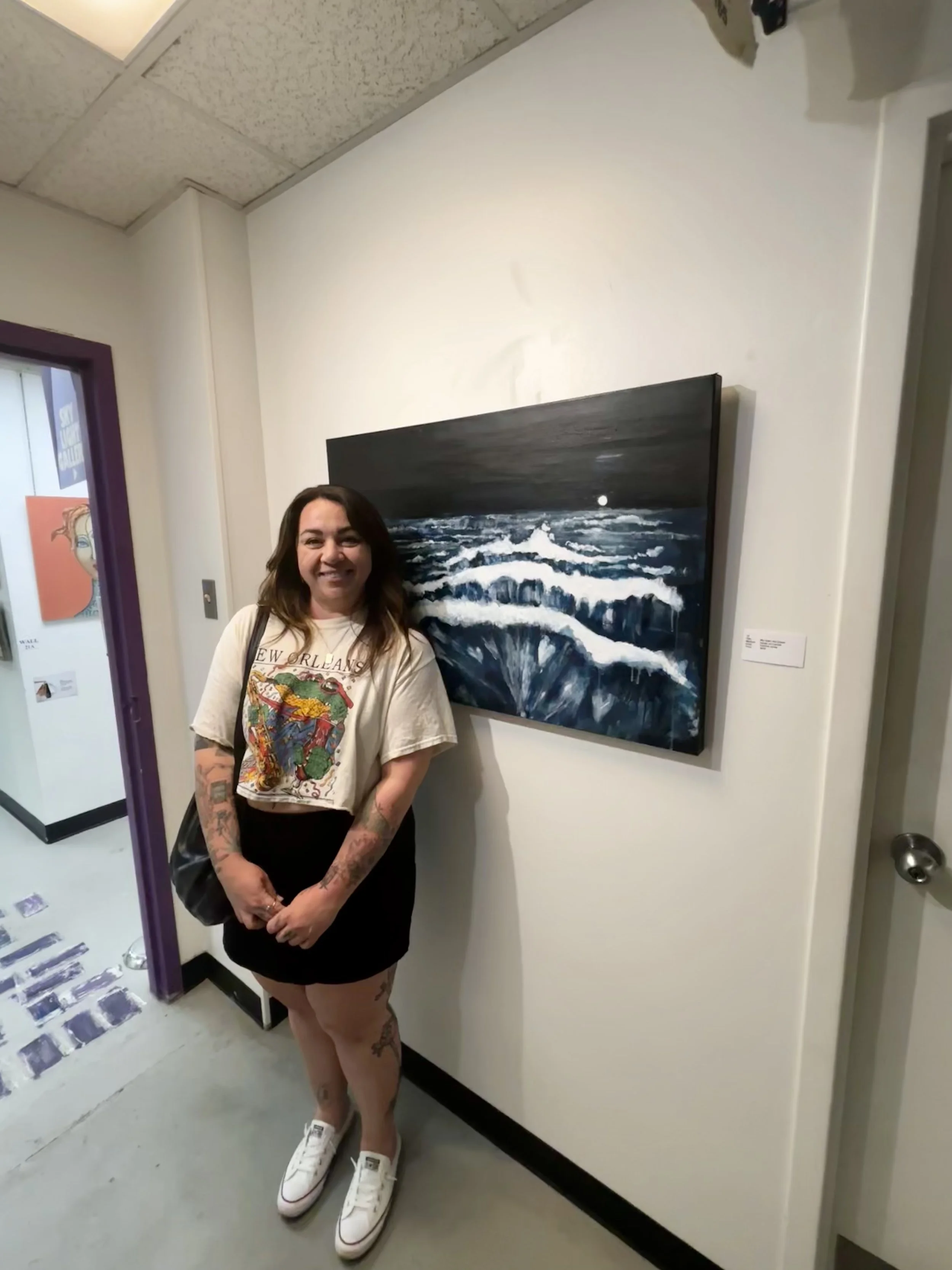 A woman with tattoos on her arms and legs, smiling, standing in an art gallery near a painting of ocean waves at night, wearing a graphic t-shirt, black skirt, and white sneakers.