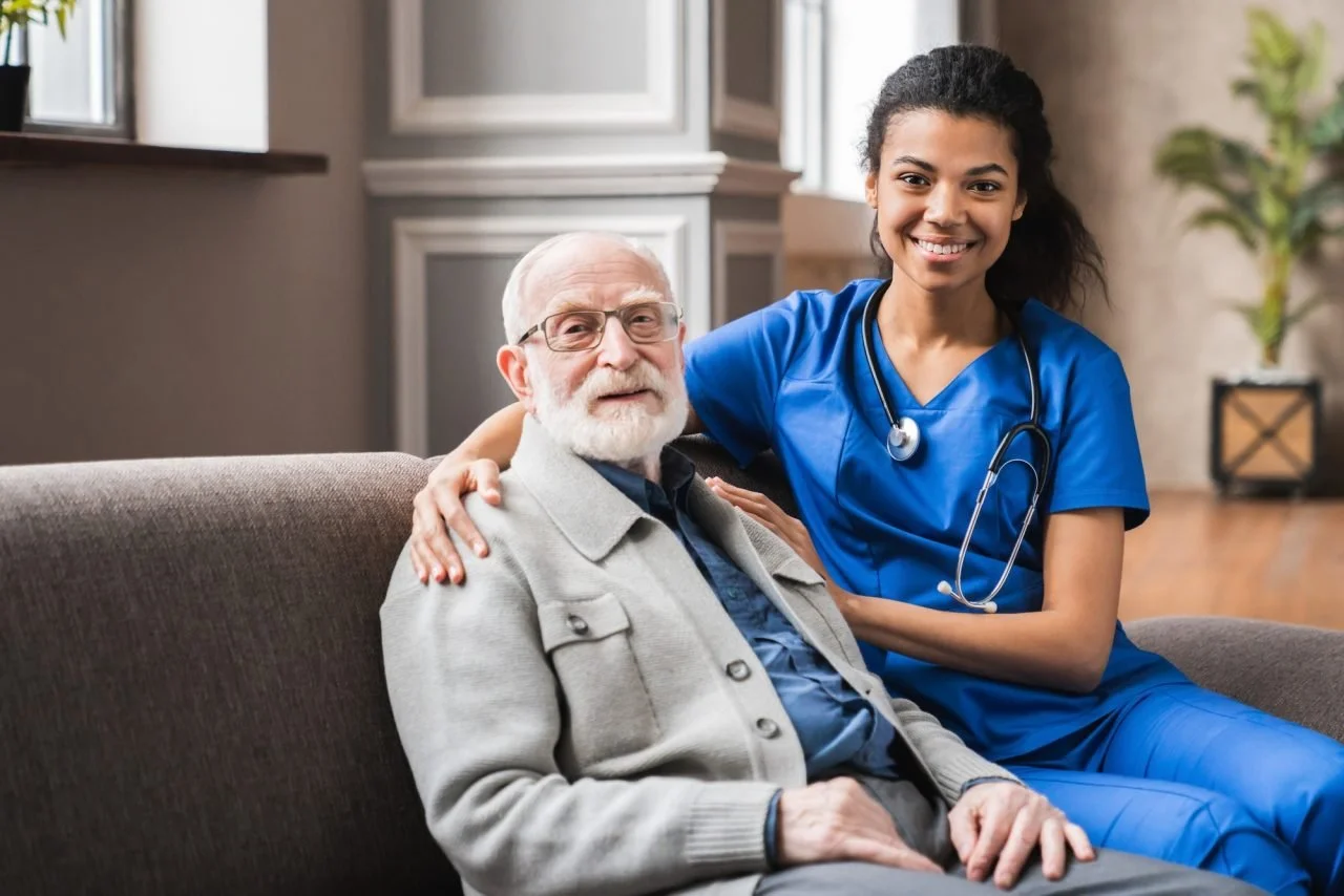 A young female healthcare worker in blue scrubs and a stethoscope with an elderly man sitting on a sofa, smiling and smiling together in a living room.