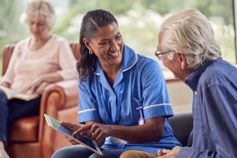 A nurse is smiling and talking to an elderly man, holding a tablet device, while an elderly woman sits on a sofa in the background.