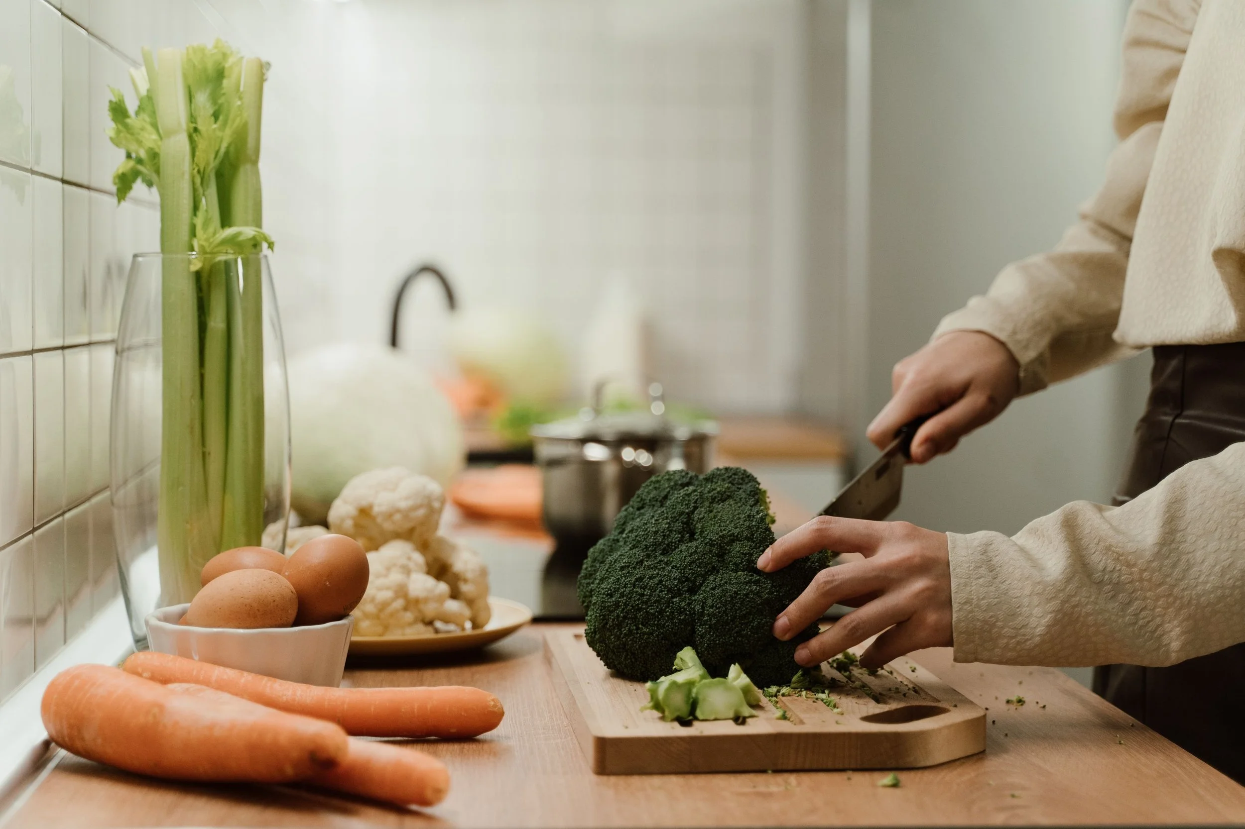Person chopping broccoli on a kitchen cutting board beside carrots, celery, eggs, cauliflower, and other fresh vegetables