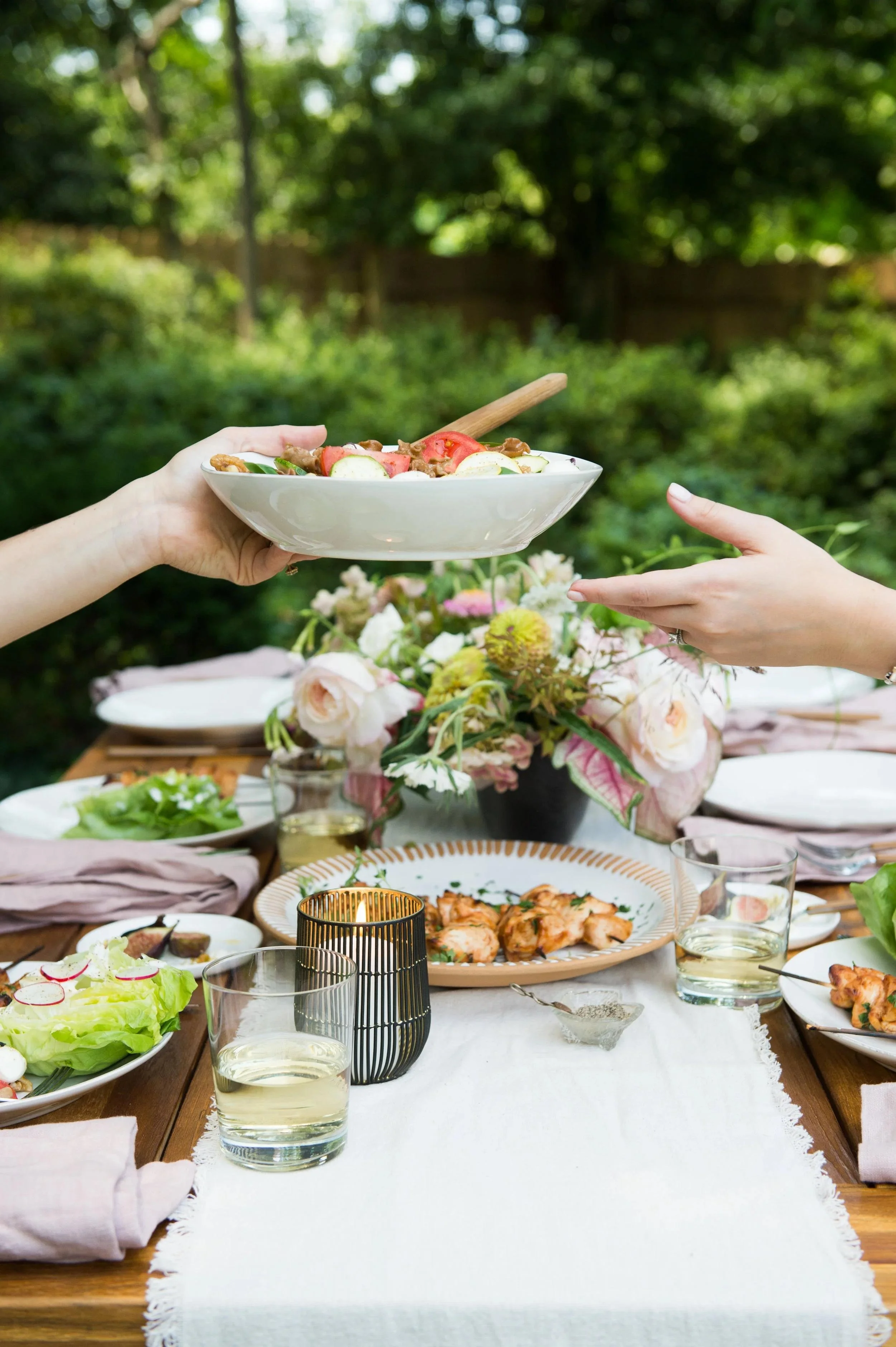 A table set for outdoor meal with dishes, glasses, and flowers, two hands reaching to place a bowl of salad on the table, lush green trees in the background.