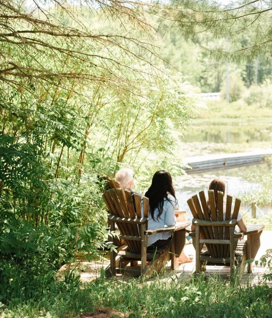Three people sitting on Adirondack chairs outdoors near a pond, surrounded by lush green trees and foliage.