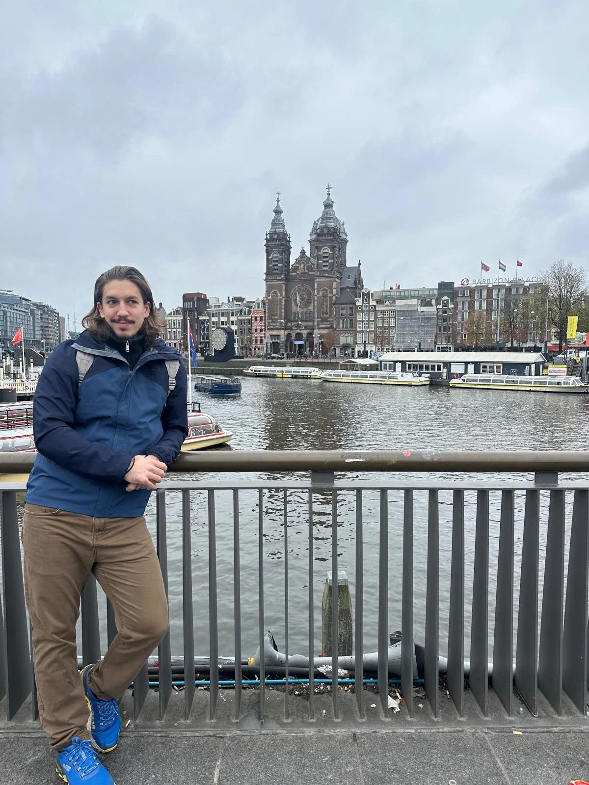 A man in a blue jacket and brown pants standing by a river with a cityscape and church in the background.