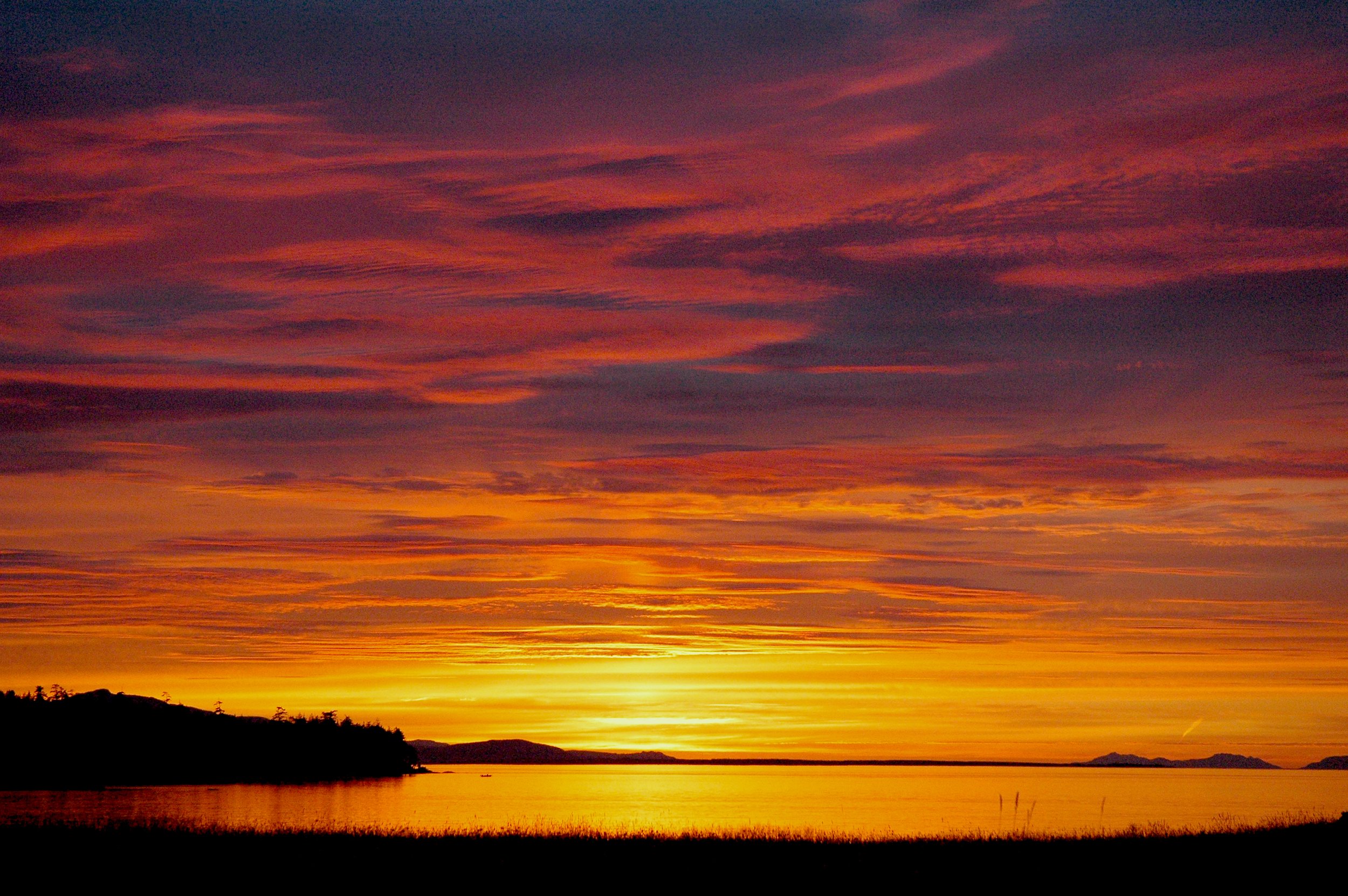 Sunset over a body of water with a silhouette of trees and hills, vibrant orange, pink, and purple clouds.