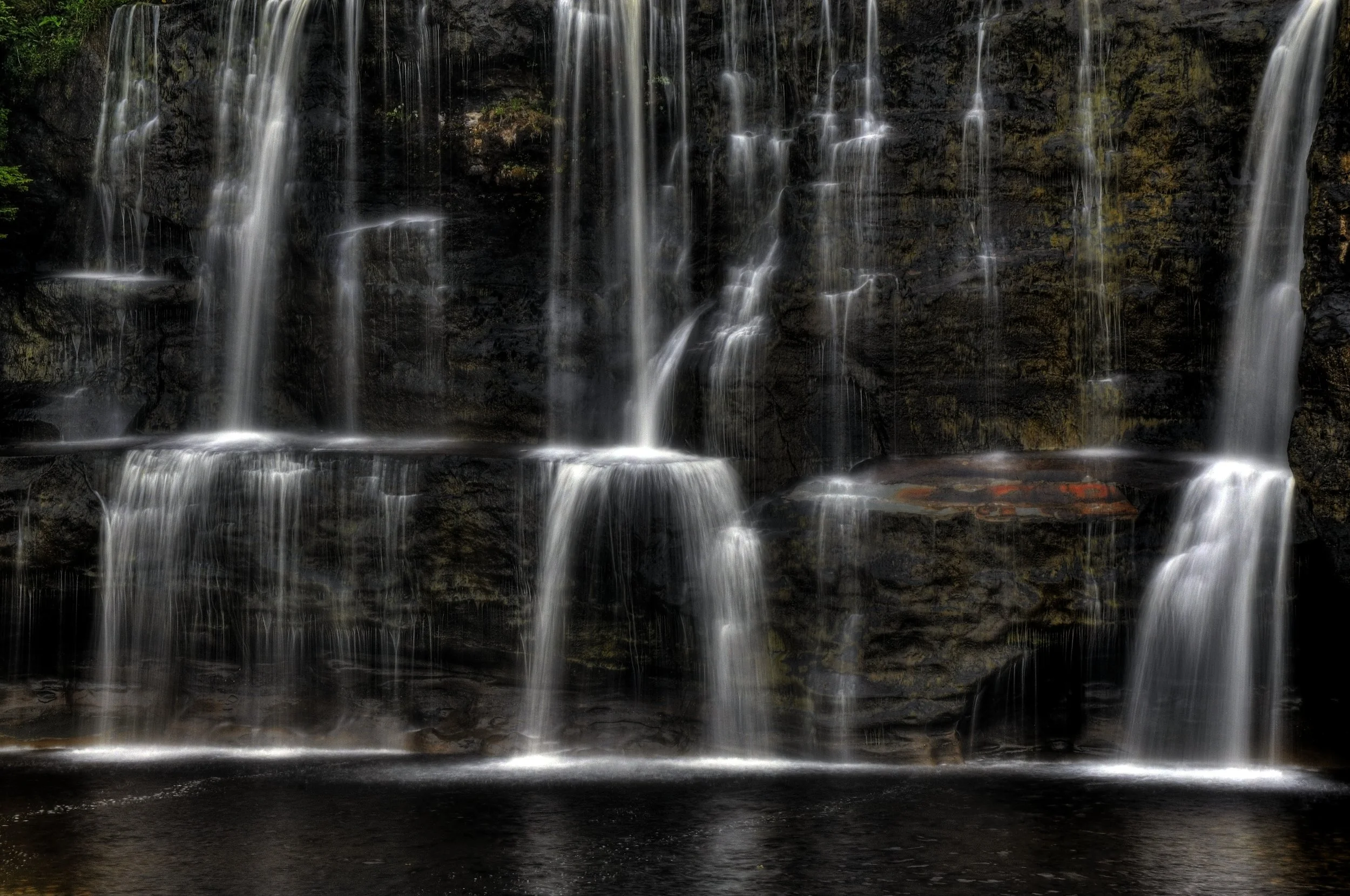 A waterfall with multiple streams flowing over a dark, moss-covered rock face into a pool below.