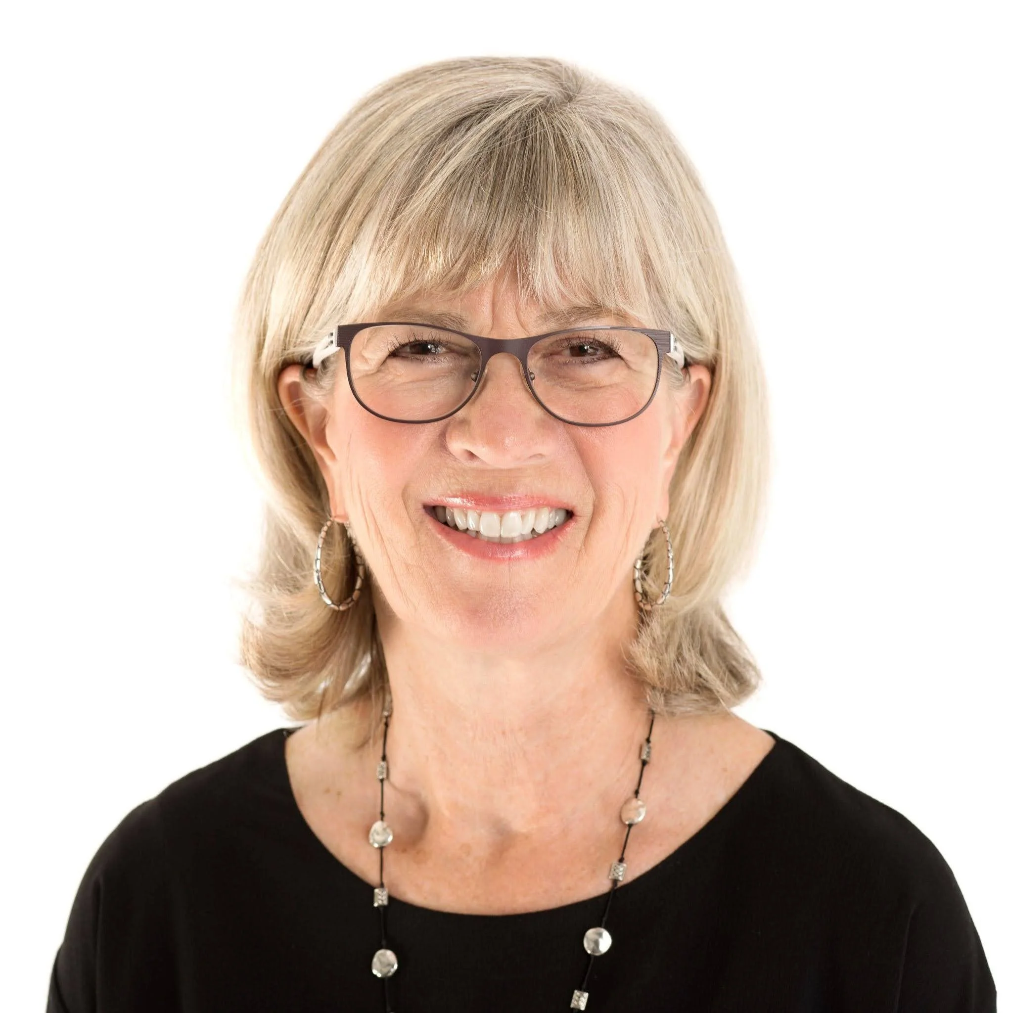 A woman with blonde hair styled in layers, wearing glasses, a black top, silver jewelry, and smiling with visible teeth.