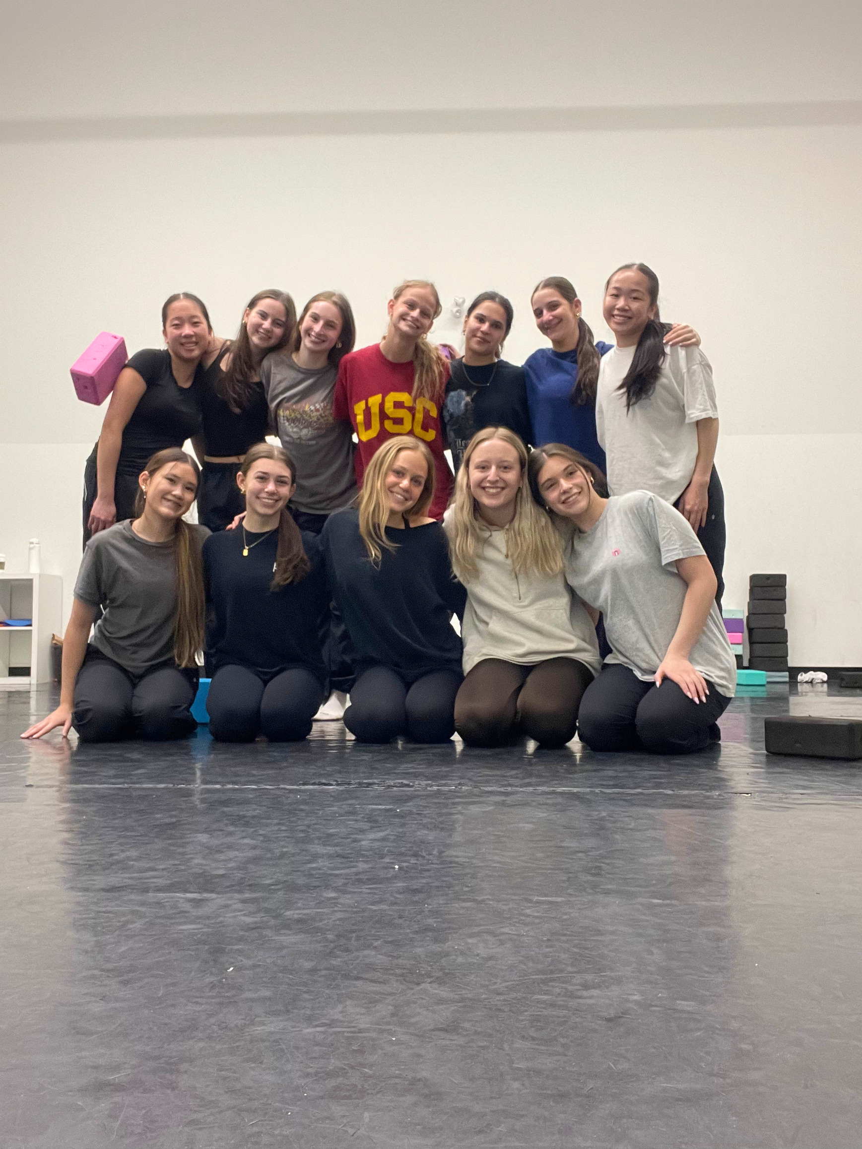 Group of 13 young women in a dance studio, some standing and some kneeling, posing for a photo with a plain white wall and yoga blocks stacked on a black floor in the background.