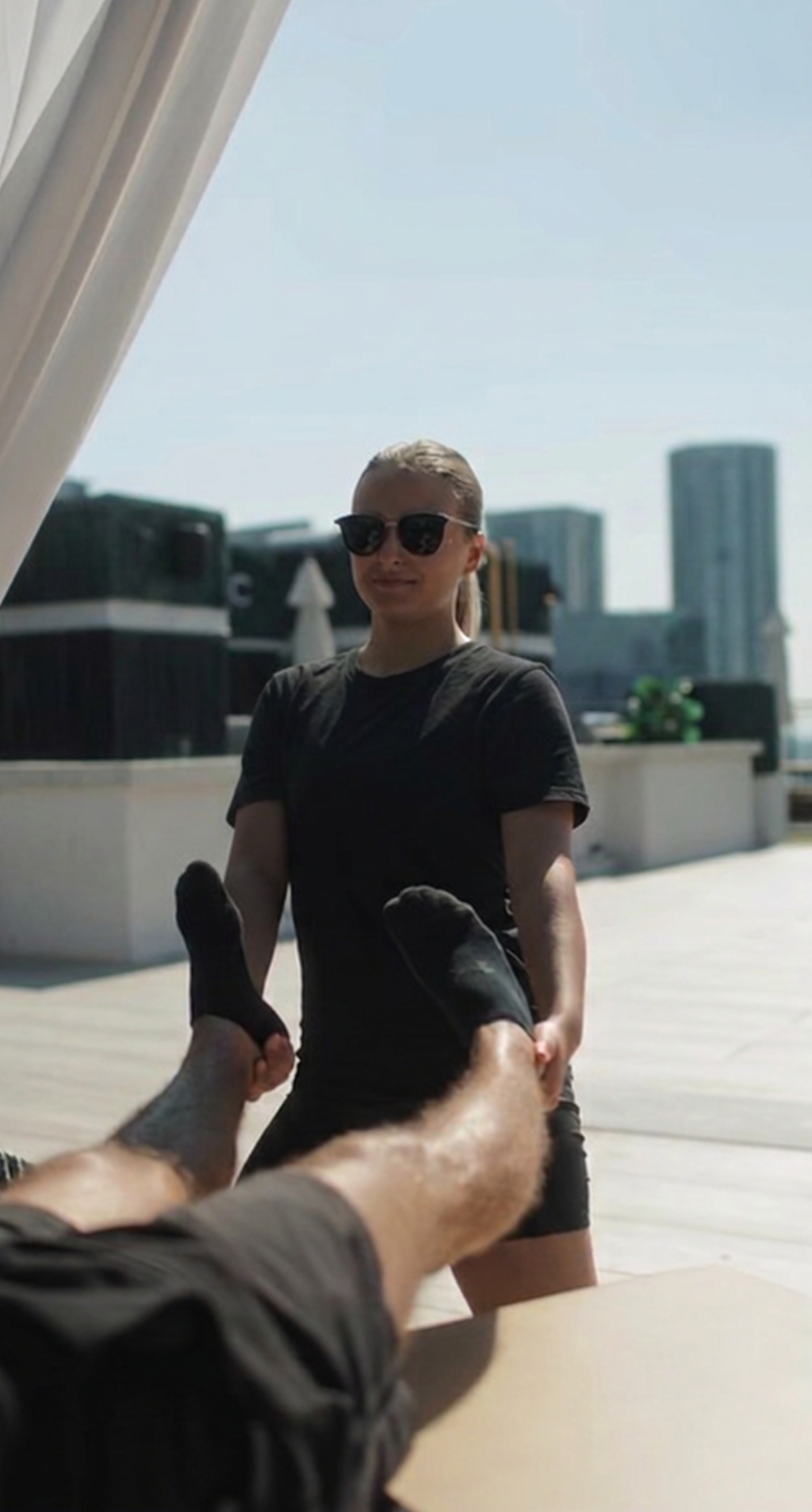 A woman standing outdoors on a rooftop with city buildings in the background, holding a person's legs and feet in her hands during daytime.