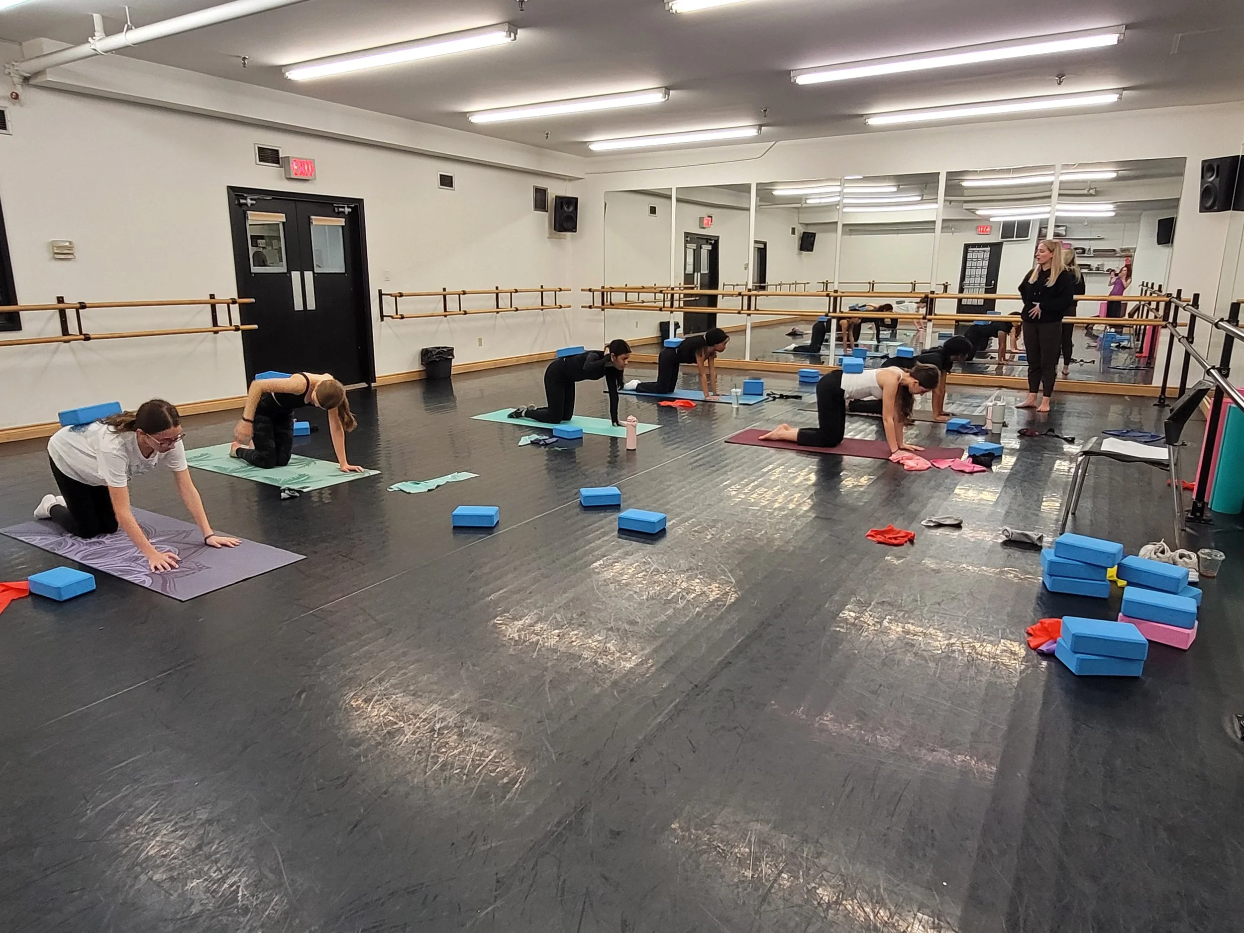 A group of people participating in a dance conditioning class in a studio with mirrors, black flooring, and barre rails. They are on yoga mats and using blue foam blocks, with an instructor observing.